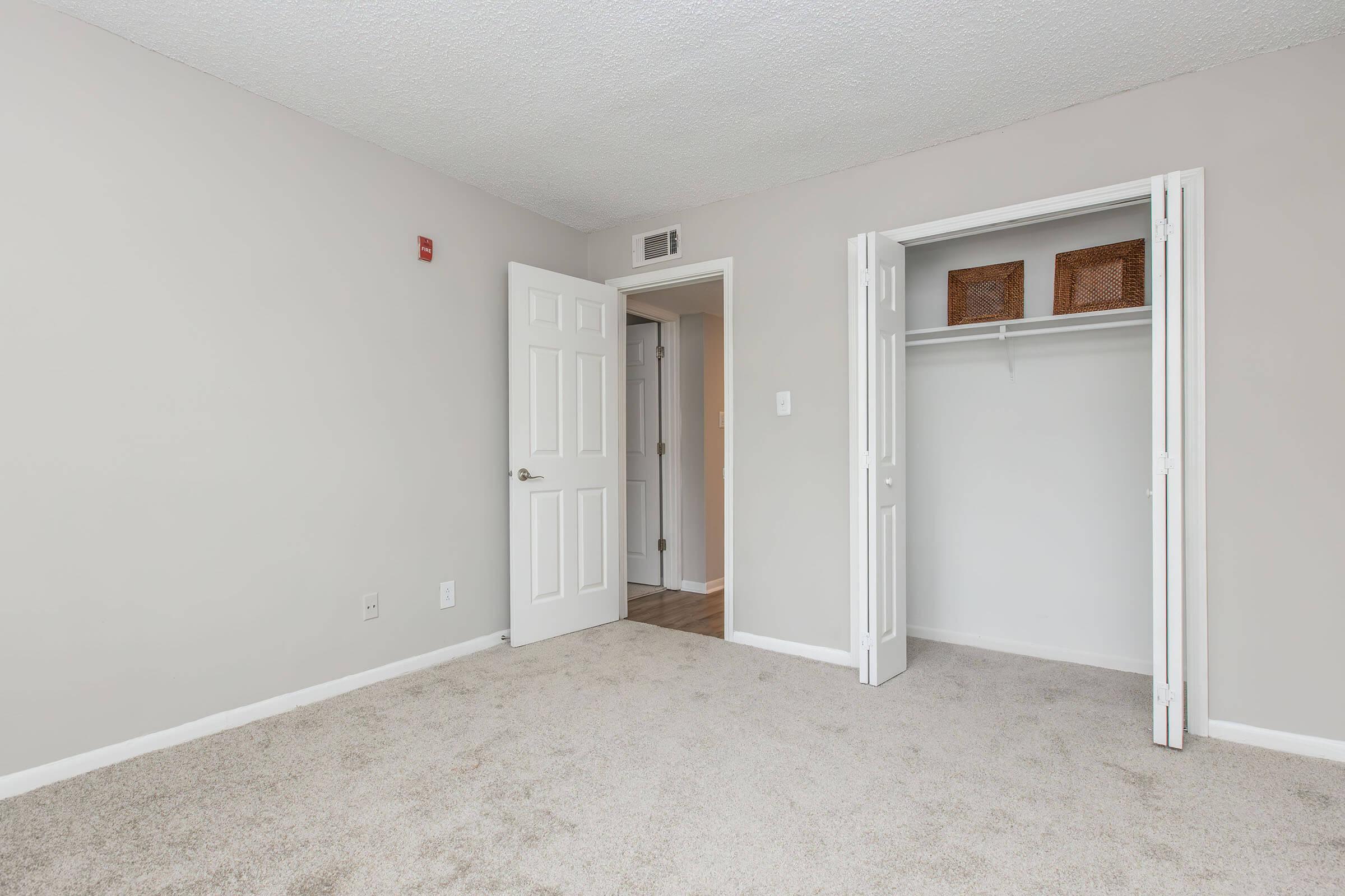 Empty bedroom with light gray walls and carpeted floor. A white closed door leads to a hallway, while an open closet with two wicker baskets is visible. The room is well-lit with no furniture.