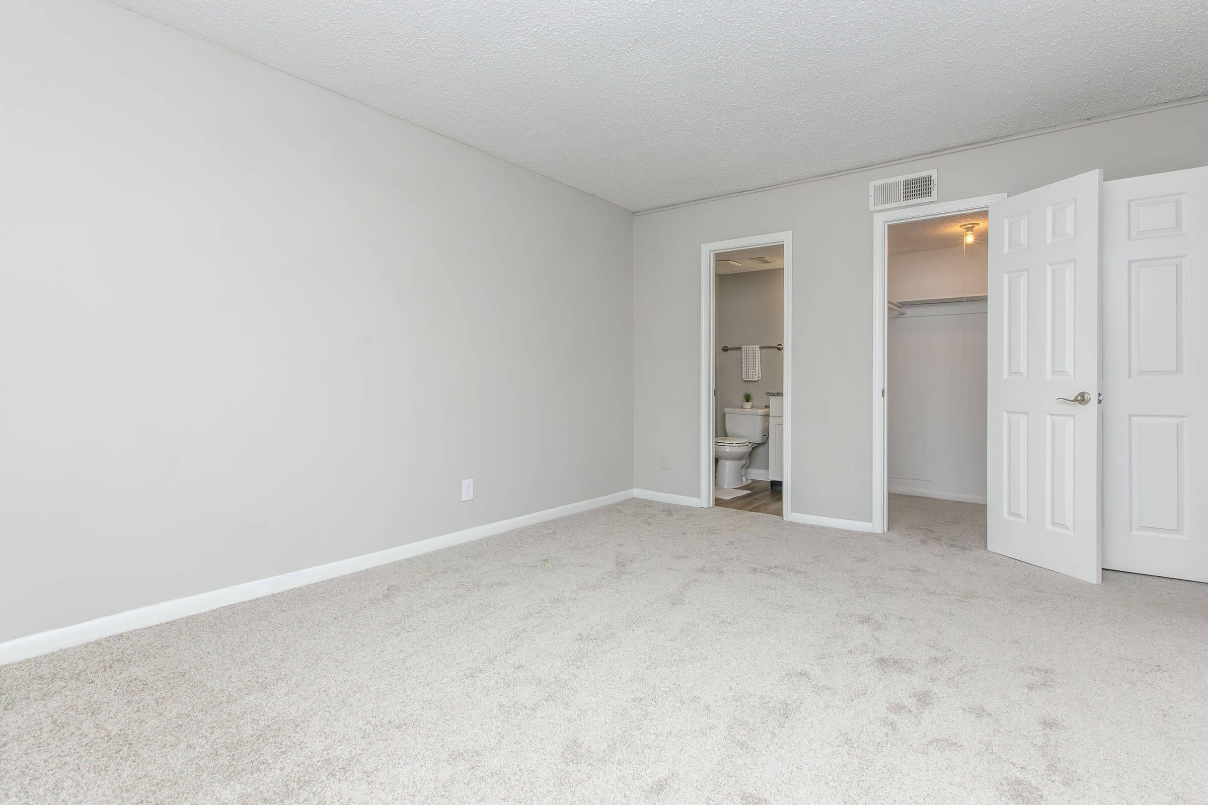 A spacious, light-colored bedroom featuring beige carpet and plain gray walls. The room has an open door leading to a small bathroom area with a toilet visible. Another door provides access to a closet. The overall ambiance is clean and minimalistic, ideal for a simple living space.
