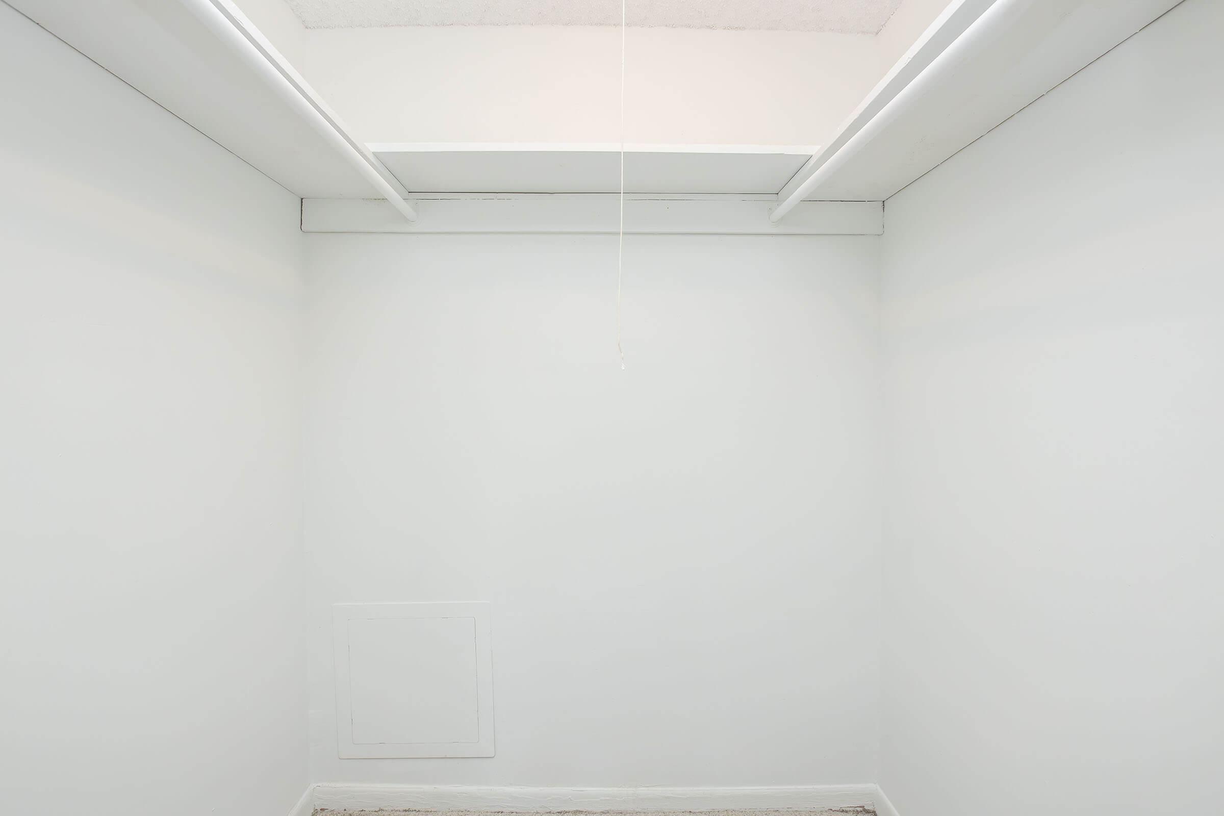 A minimalistic, empty room with white walls and a ceiling visible. There is a small access panel on the lower left side of the wall, and a single light fixture hanging from the ceiling. The floor is bare and the overall space feels open and uncluttered.