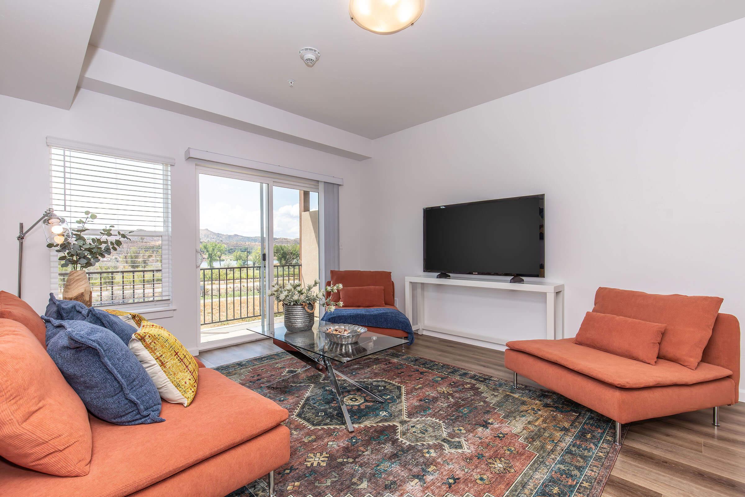 A modern living room featuring two orange sofas, a glass coffee table on a patterned rug, and a large TV mounted on a white console. There are large windows allowing natural light, offering a view of the outdoors. Decorative pillows and a floor lamp add a warm touch to the space.
