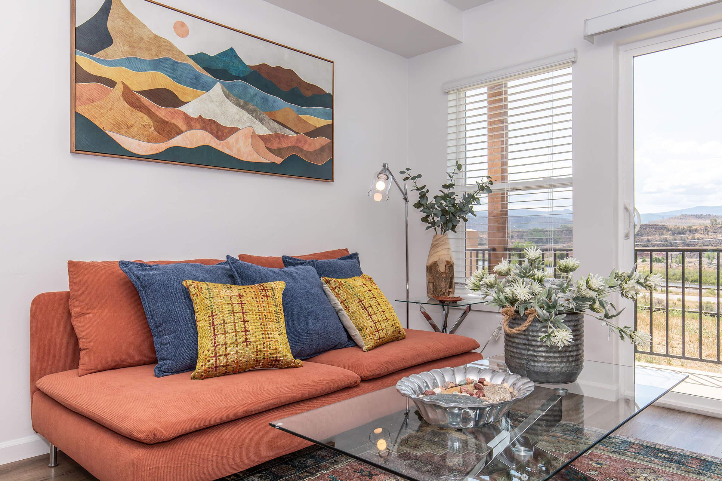 A cozy living room featuring an orange sectional sofa with colorful pillows. A modern artwork depicting mountains hangs above the sofa. There's a glass coffee table with a decorative bowl on it, and a vase of flowers nearby. Natural light floods in through a window, creating a warm and inviting atmosphere.