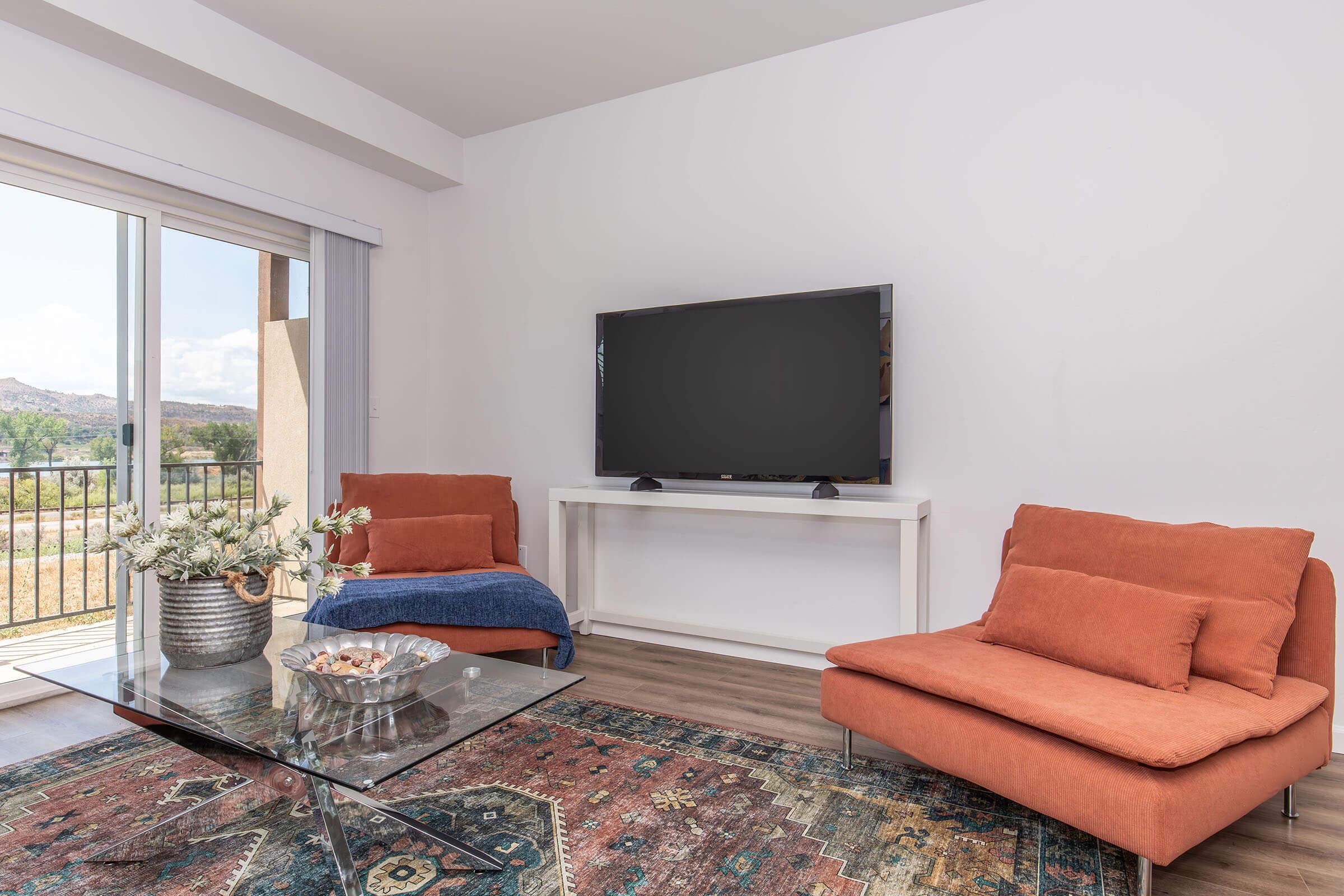 A modern living room featuring two orange armchairs and a glass coffee table on a colorful area rug. A large flat-screen TV is mounted on the wall above a white console table. Natural light streams in from a nearby balcony, adding warmth to the space with a scenic view outside.