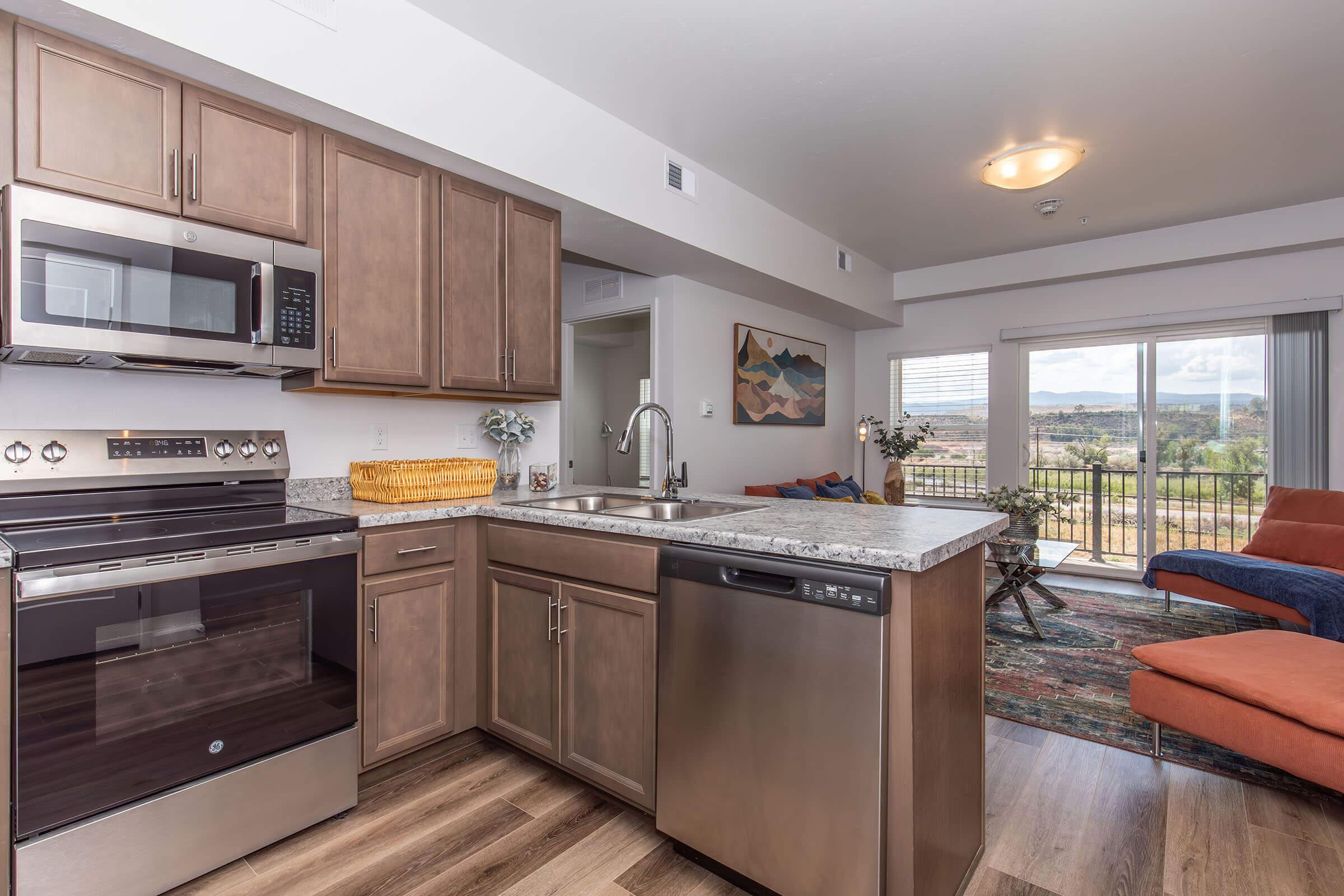 Modern kitchen featuring stainless steel appliances, including a microwave and oven, complemented by wooden cabinets and granite countertops. A dining area with a view of mountains can be seen through a sliding glass door, along with a cozy living space furnished with a blue couch and a colorful rug.