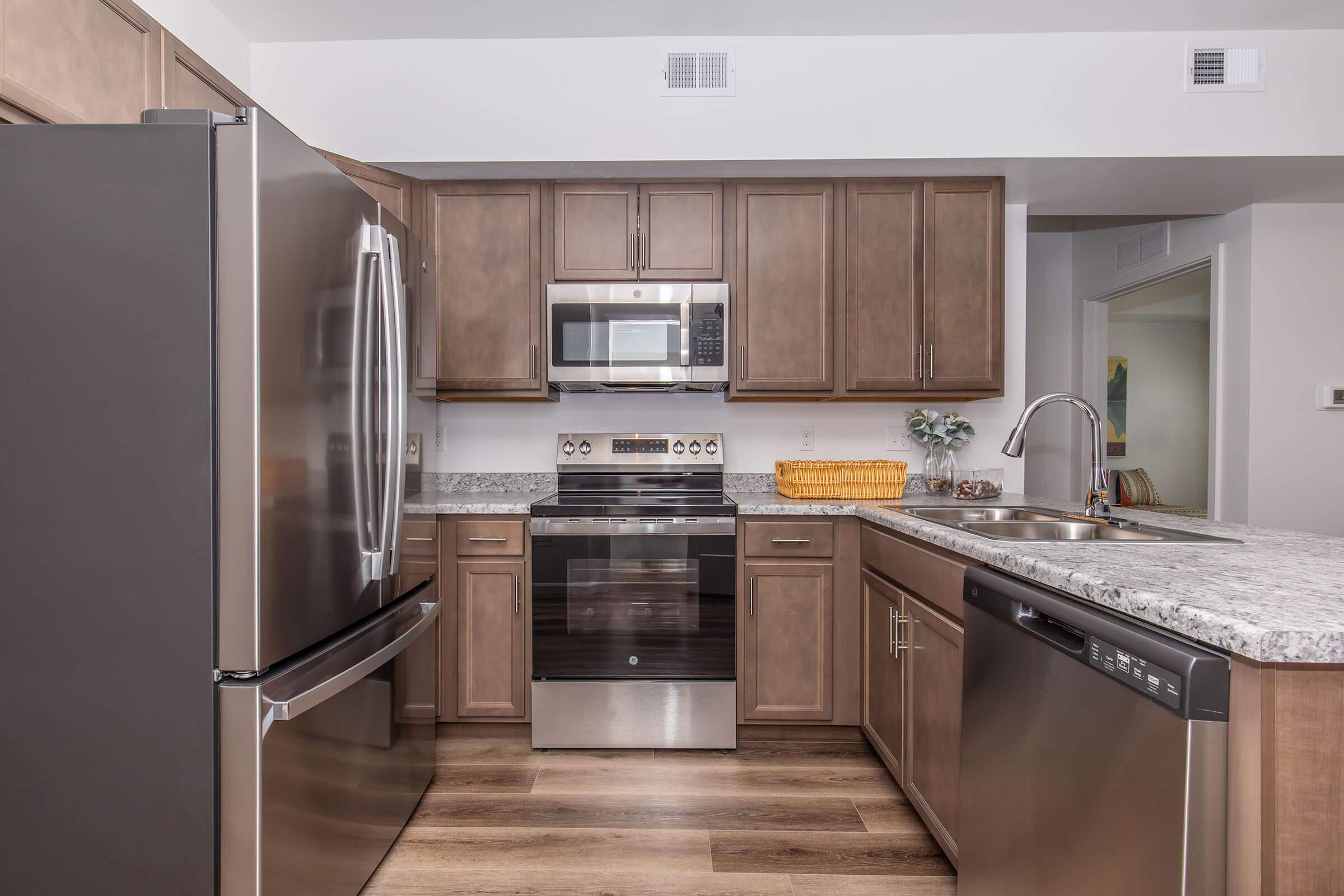 Modern kitchen featuring stainless steel appliances, including a refrigerator, oven, and microwave. The cabinets are a rich brown wood, and the countertops are a light granite. A stainless steel sink is positioned near the dishwasher, and a woven basket adds a decorative touch on the counter.