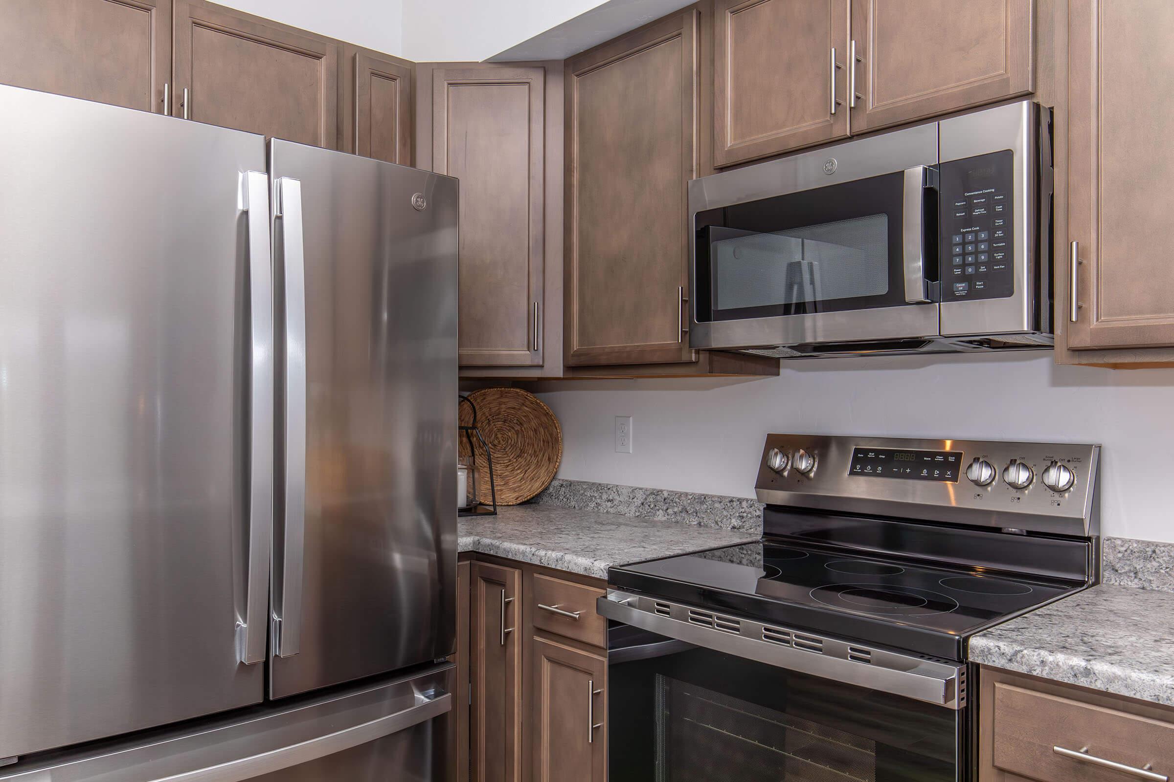 A modern kitchen featuring stainless steel appliances, including a refrigerator, microwave, and oven. The cabinets are a warm brown, and the countertops are a light gray granite. The layout is sleek and contemporary, emphasizing a clean and functional design.