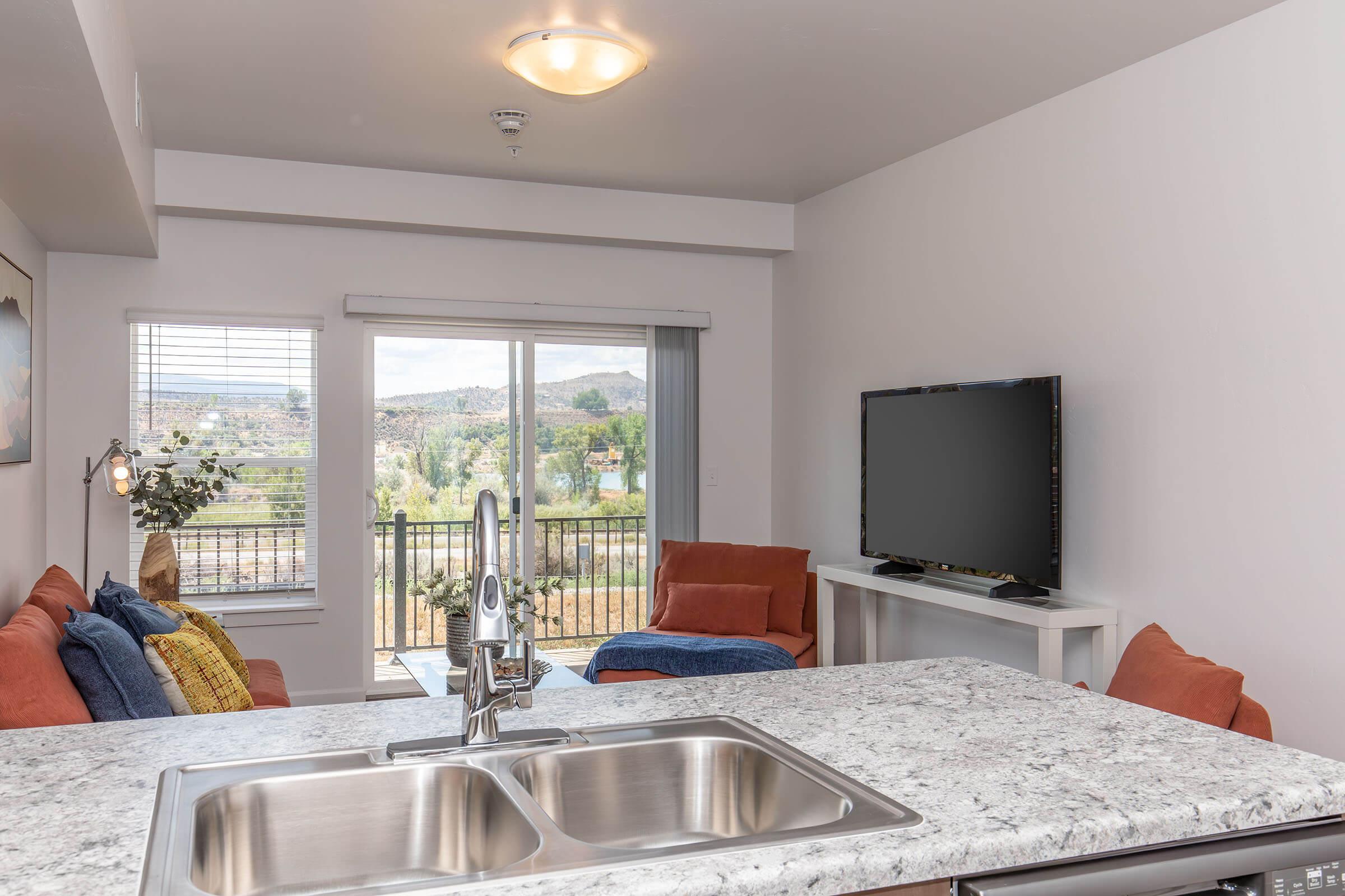 A cozy living room featuring a granite kitchen island. The space includes two orange sofas, a television on a white stand, and large windows with a scenic view of mountains and greenery. Bright natural light enhances the inviting atmosphere.