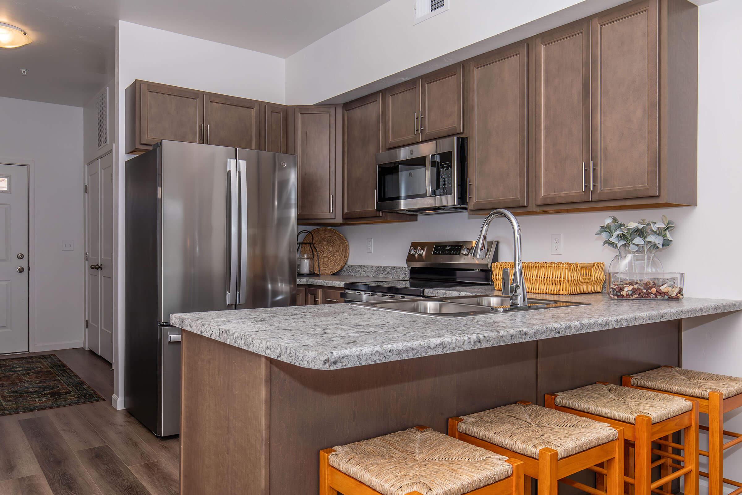 A modern kitchen featuring gray cabinets, stainless steel appliances, a large granite countertop, and three wooden stools at the island. There’s a basket with decorative items on the counter and a rug on the floor, creating a warm and inviting atmosphere.