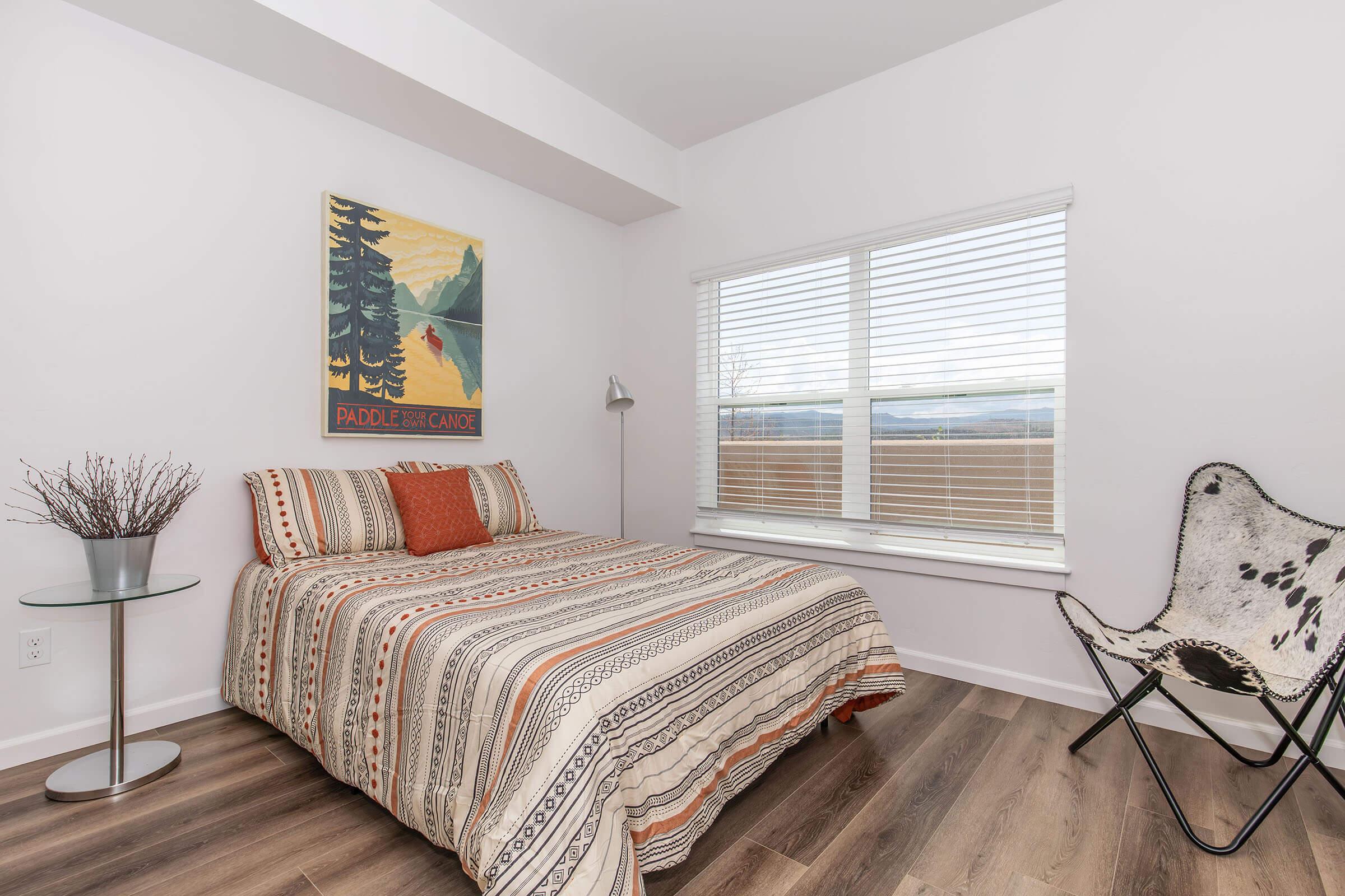 A cozy bedroom featuring a striped bed with an orange pillow, a modern writing desk with a small vase, and a unique chair. Natural light flows in through a large window with blinds, showcasing a serene view. A nature-themed poster hangs on the wall, adding character to the space.