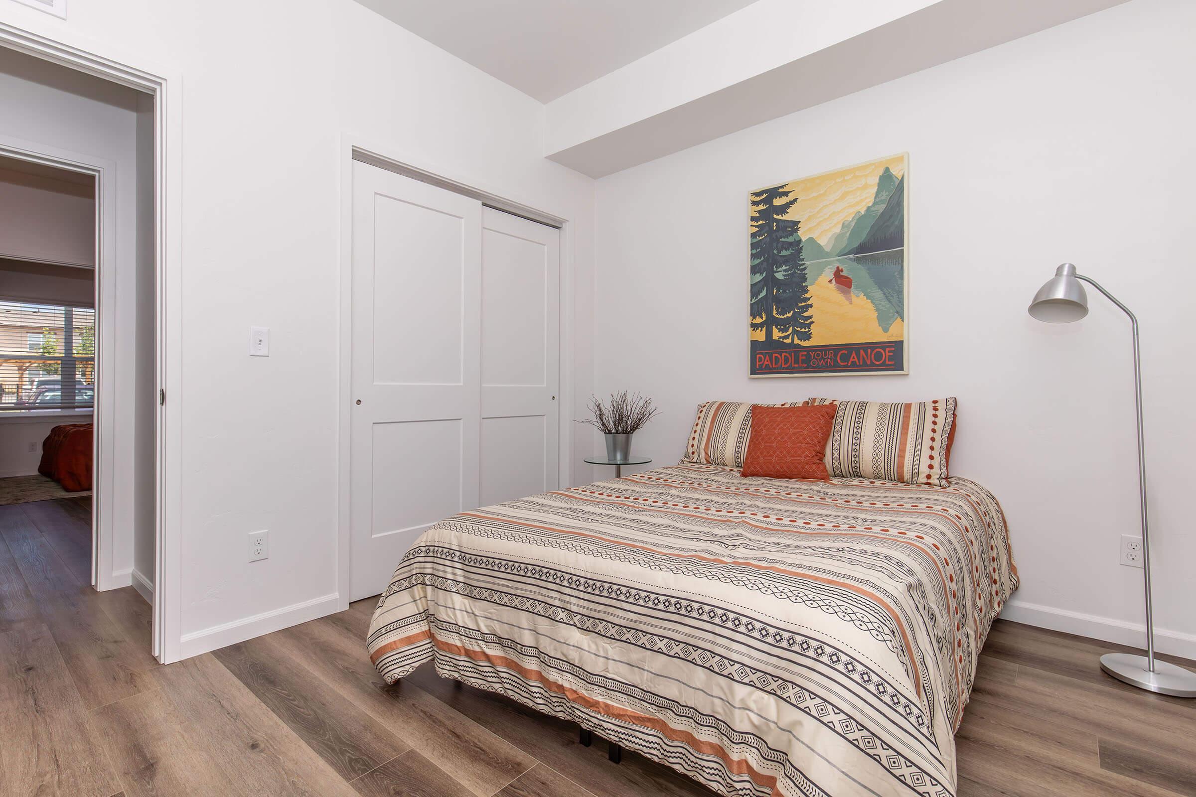 A cozy bedroom featuring a striped bedspread, a bedside table with decorative items, and a modern floor lamp. A painting of a canoe scene is displayed on the wall. The room has light-colored walls and wooden flooring, creating a warm and inviting atmosphere. A closet door is visible in the background.