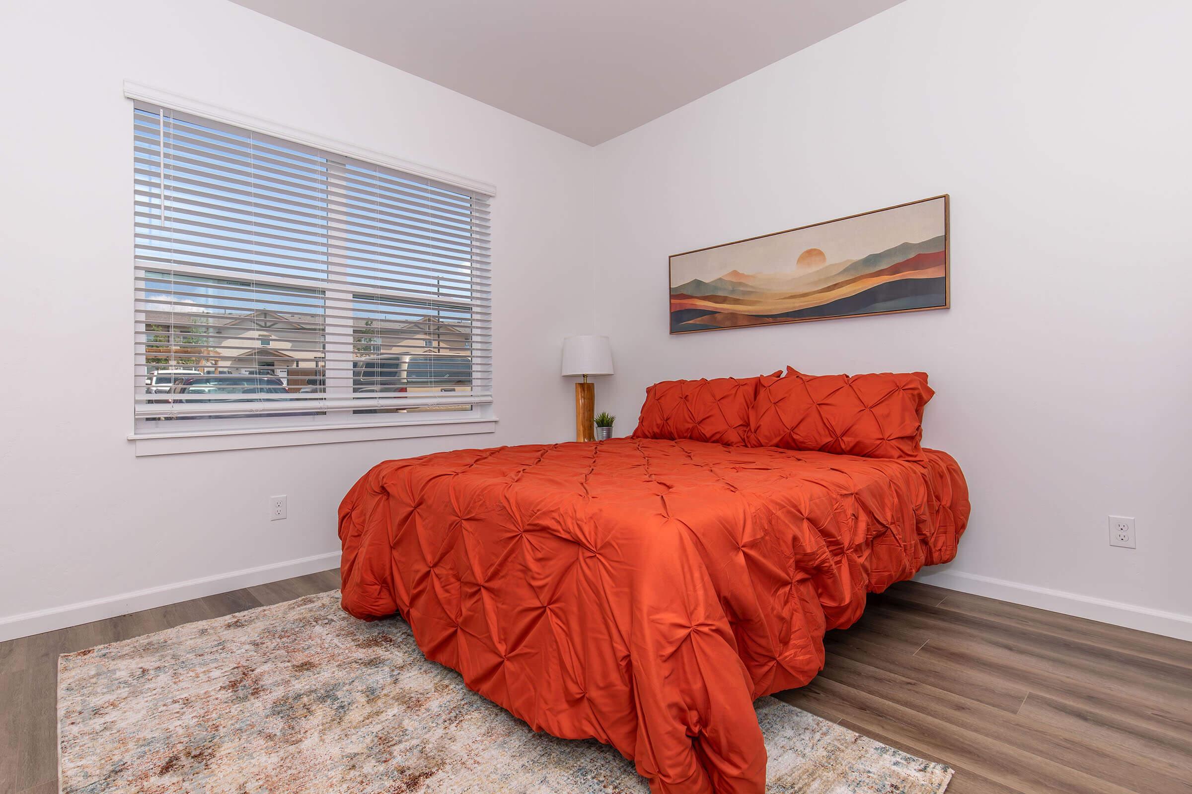 A brightly lit bedroom featuring a large bed with an orange quilted cover, a decorative pillow, and a small lamp on a bedside table. A window with white blinds allows natural light in, revealing a view of the outdoors. The floor is covered with a soft area rug, and the walls are painted white.