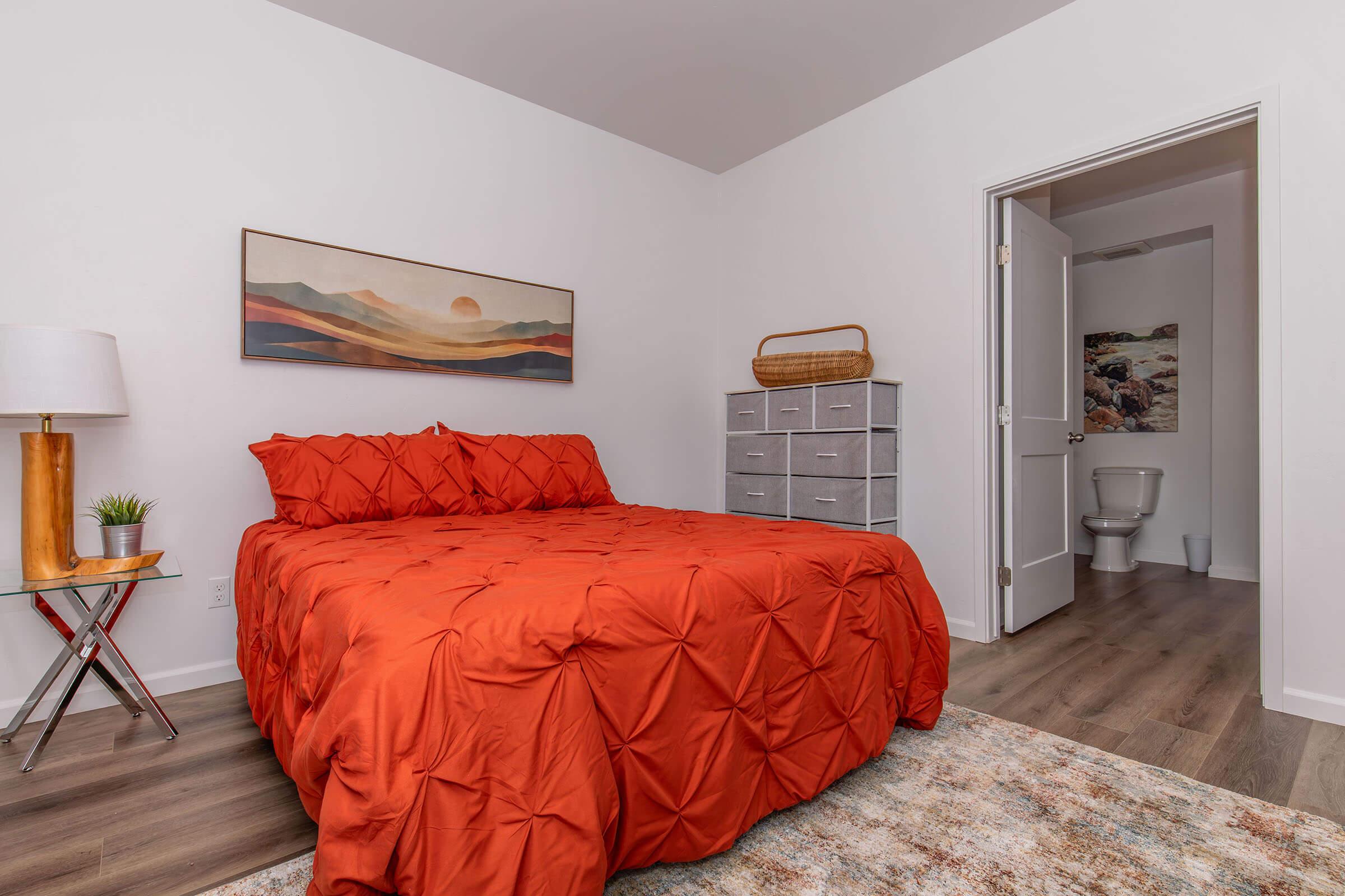 A cozy bedroom featuring a bright orange quilted bedspread, a small bedside lamp, a gray dresser, and an artwork above the bed. A door leads to a bathroom with a toilet visible. The room has wooden flooring and a decorative area rug, creating a warm and inviting atmosphere.