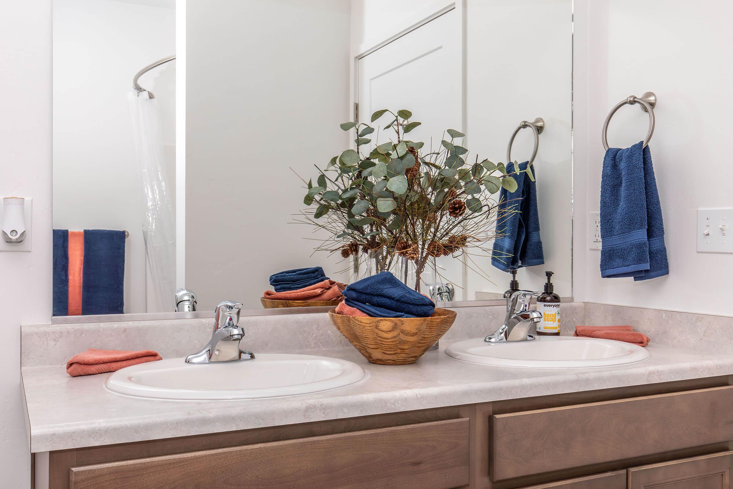 A modern bathroom with double sinks and a light-colored countertop. A decorative arrangement of eucalyptus branches sits in a basket between the sinks. Blue hand towels hang from a wall-mounted rack, and matching towels are piled next to a bottle of hand soap. A shower curtain is visible in the background.