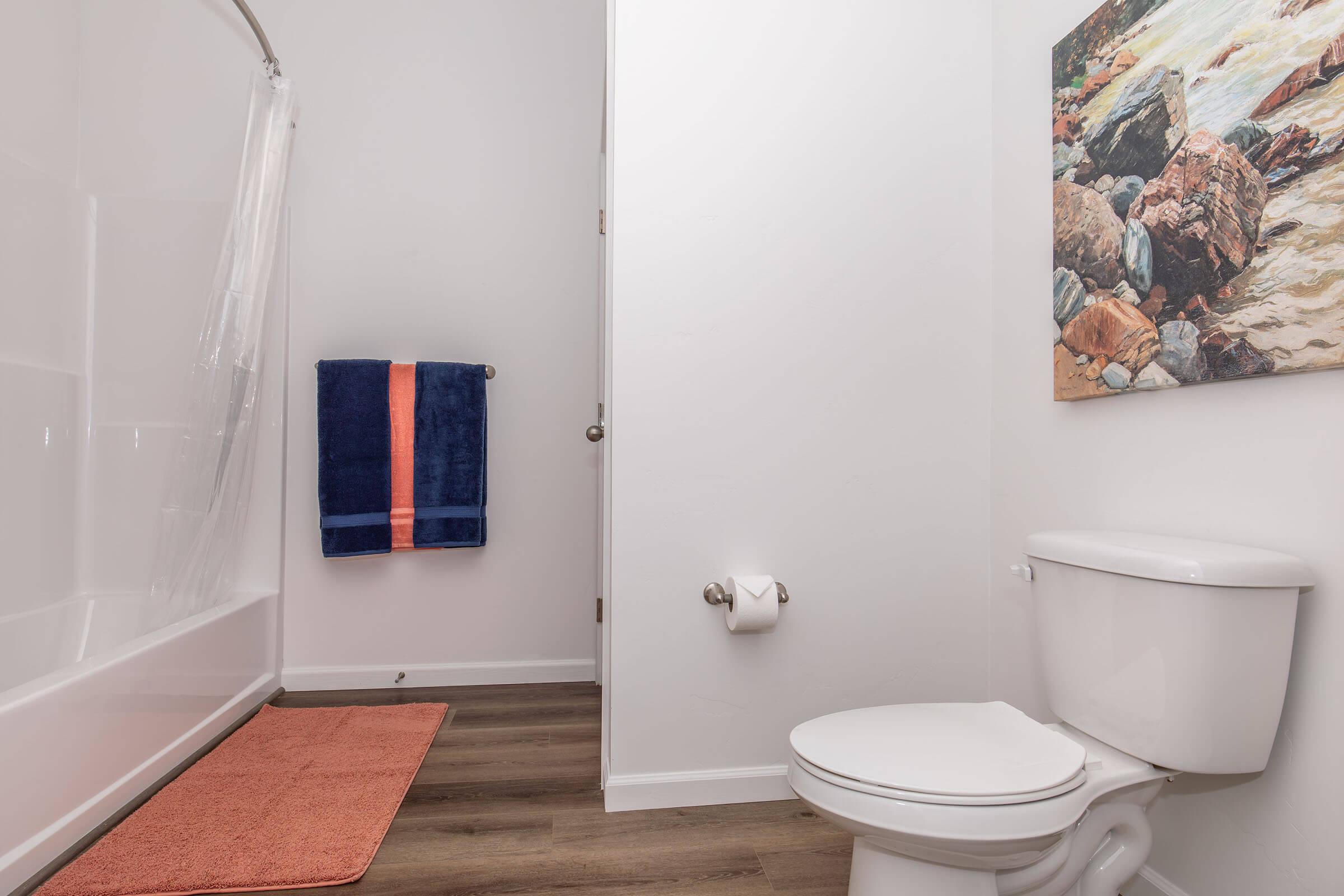 A clean bathroom featuring a bathtub with a shower curtain, a toilet, and a towel rack with a blue towel. The floor is covered with an orange mat, and a landscape artwork hangs on the wall. The overall decor is modern and tidy, with light-colored walls and wooden flooring.