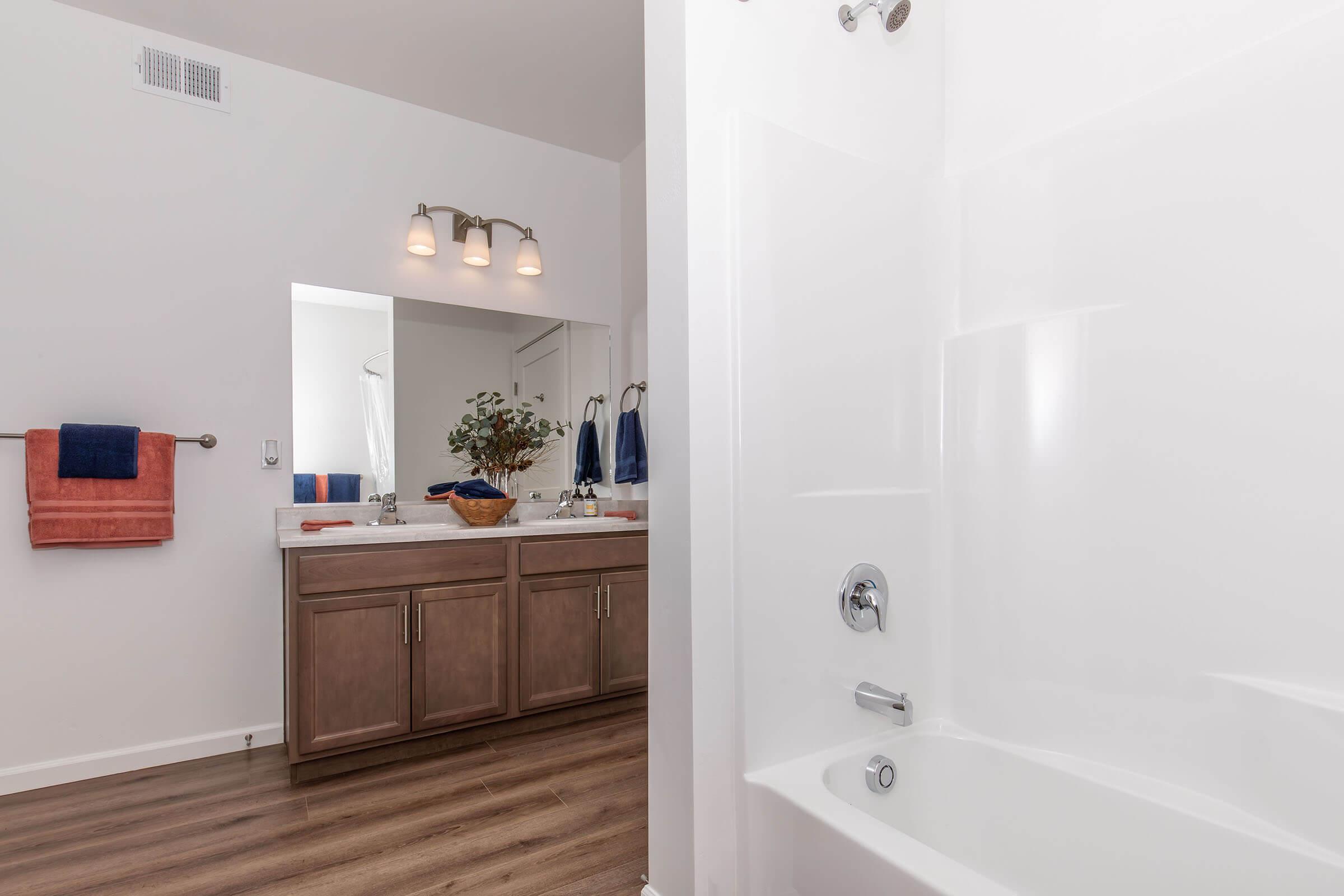 A modern bathroom featuring a bathtub on the right and a vanity with two sinks on the left. The vanity has a large mirror and is decorated with a vase of flowers. Towels in blue and orange hang nearby, with wooden flooring visible throughout the space. The walls and fixtures are bright and clean.