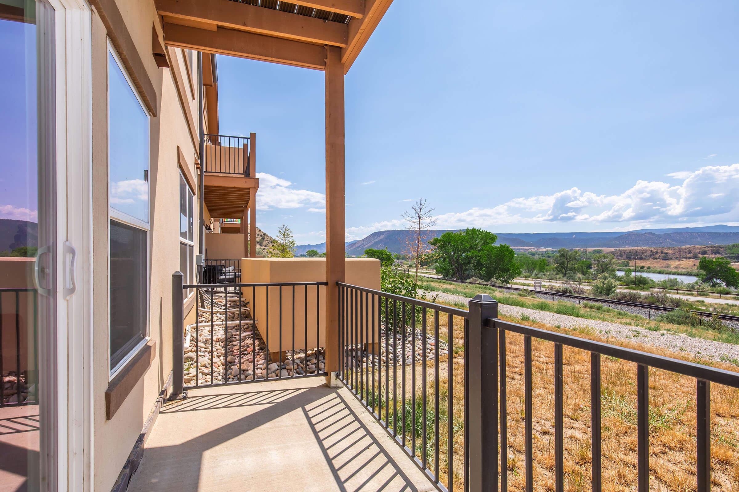 View from a balcony overlooking a scenic landscape with mountains in the background, greenery, and a river. The balcony features a railing and stone landscaping, with a clear blue sky and fluffy clouds enhancing the natural beauty of the setting.