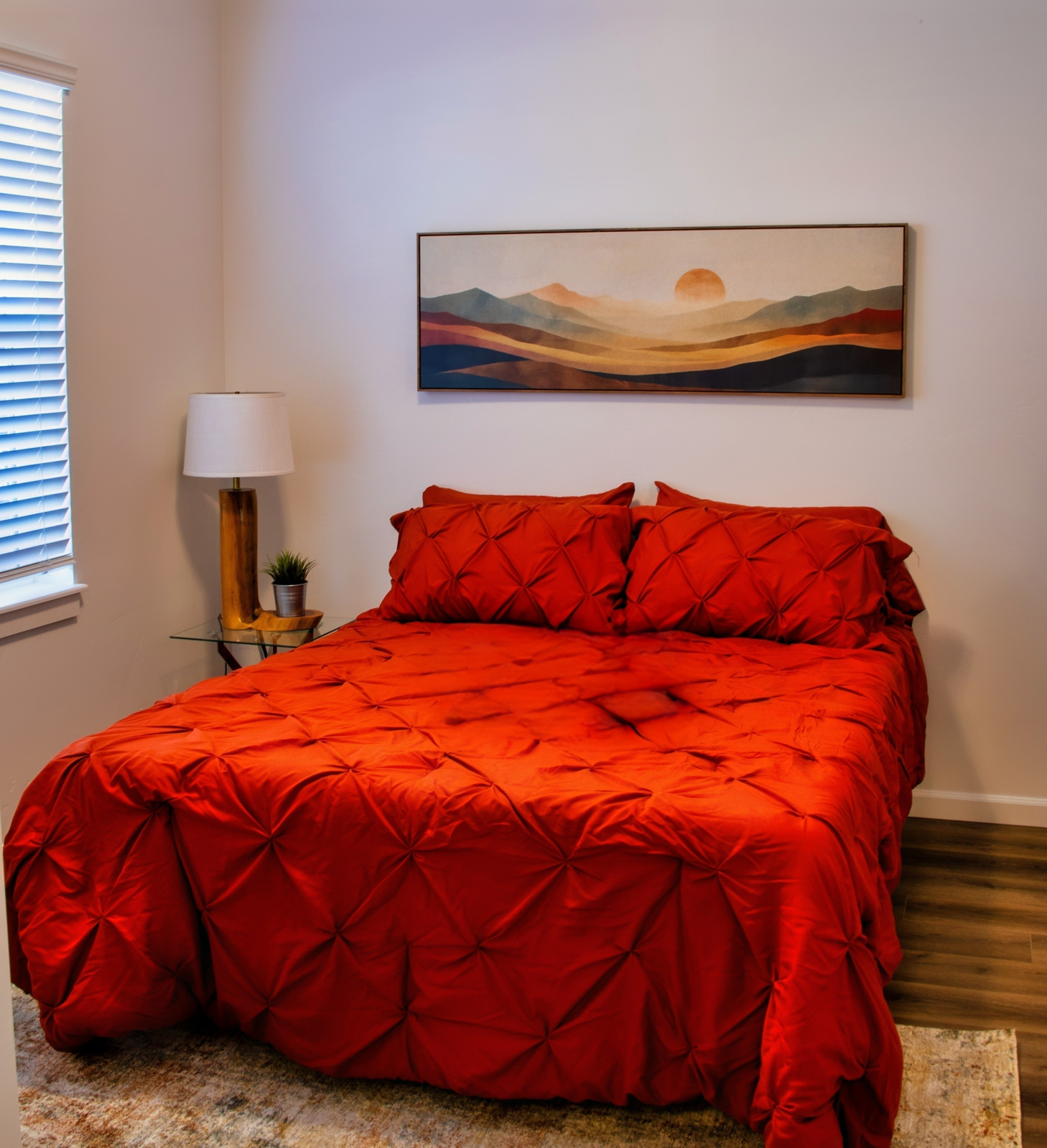 A cozy bedroom featuring a bed with a bright red comforter and matching pillows. The bed is positioned against a white wall, with a painting of a sunset over mountains above it. A small nightstand with a lamp and a potted plant is beside the bed, and there is a rug on the floor. Natural light filters in through a window with horizontal blinds.