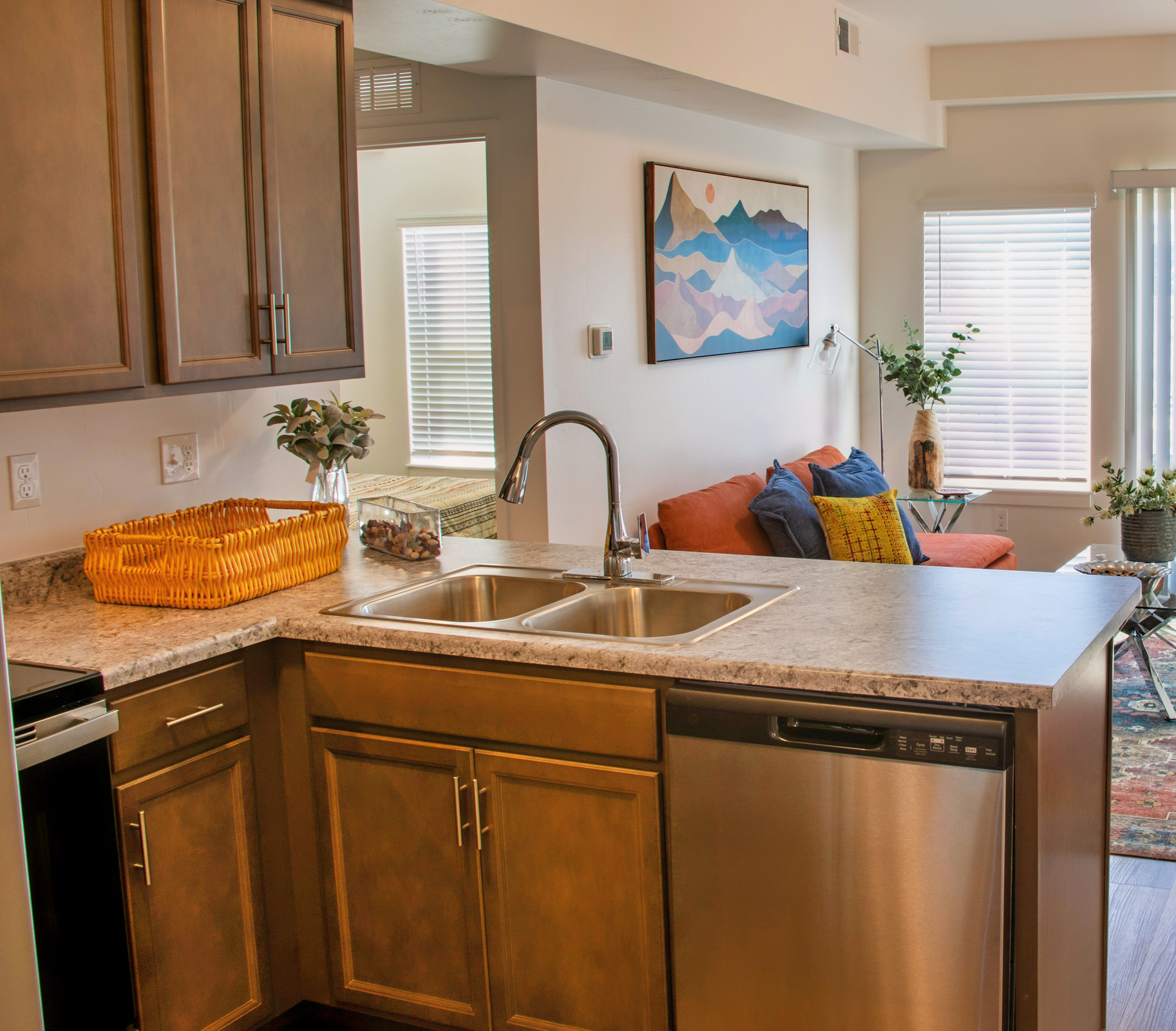 Modern kitchen with brown cabinets and a granite countertop featuring a double sink and a dishwasher. In the background, a cozy living room is visible with a colorful couch, a coffee table, and decorative plants. Natural light floods the space through large windows, creating a warm and inviting atmosphere.