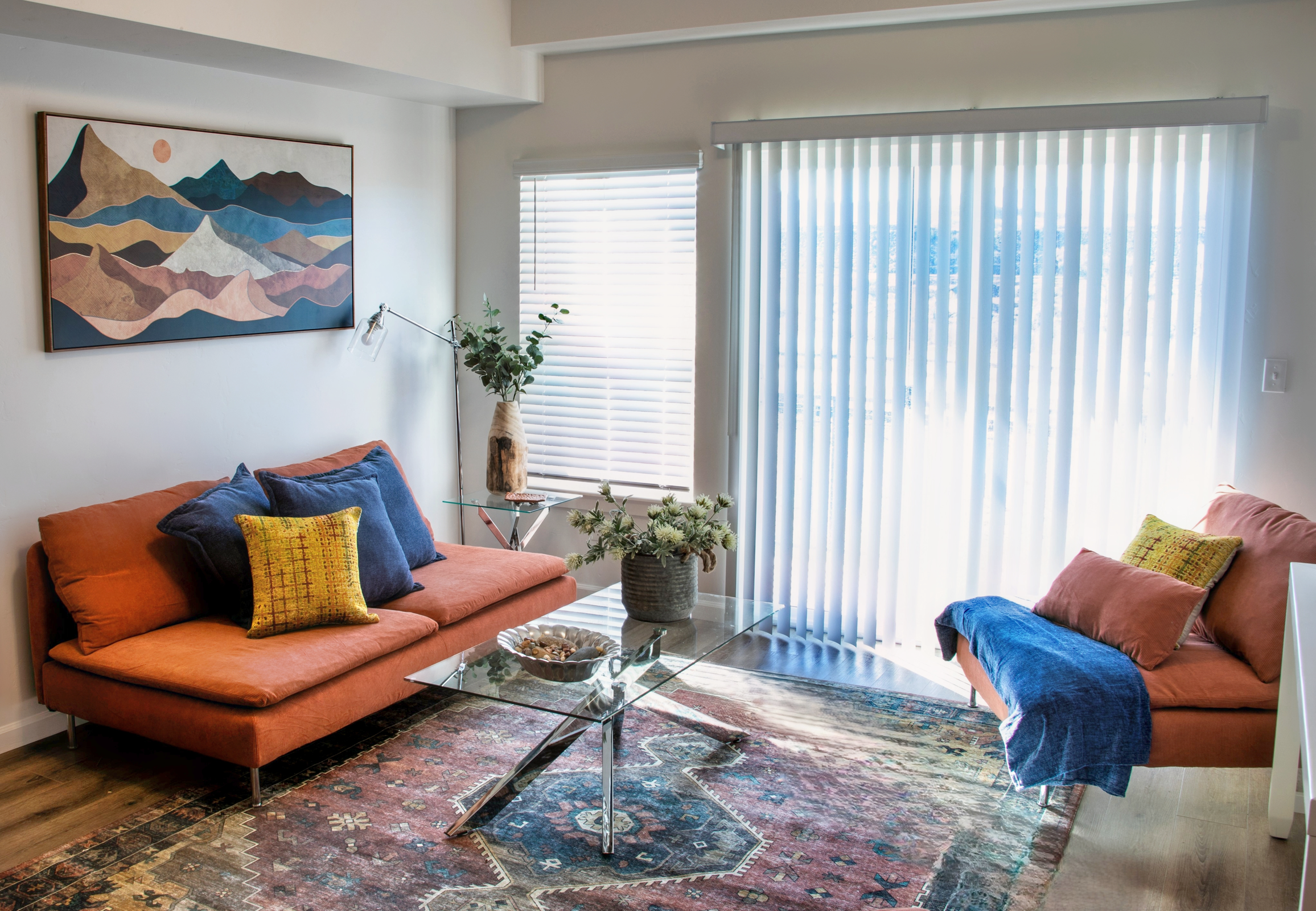 A cozy living room featuring an orange sofa and armchair with colorful cushions. A glass coffee table sits on a patterned rug. A potted plant is on the table, and a floor lamp illuminates the space. The large window has vertical blinds, allowing natural light to fill the room, with a mountain-themed artwork on the wall.