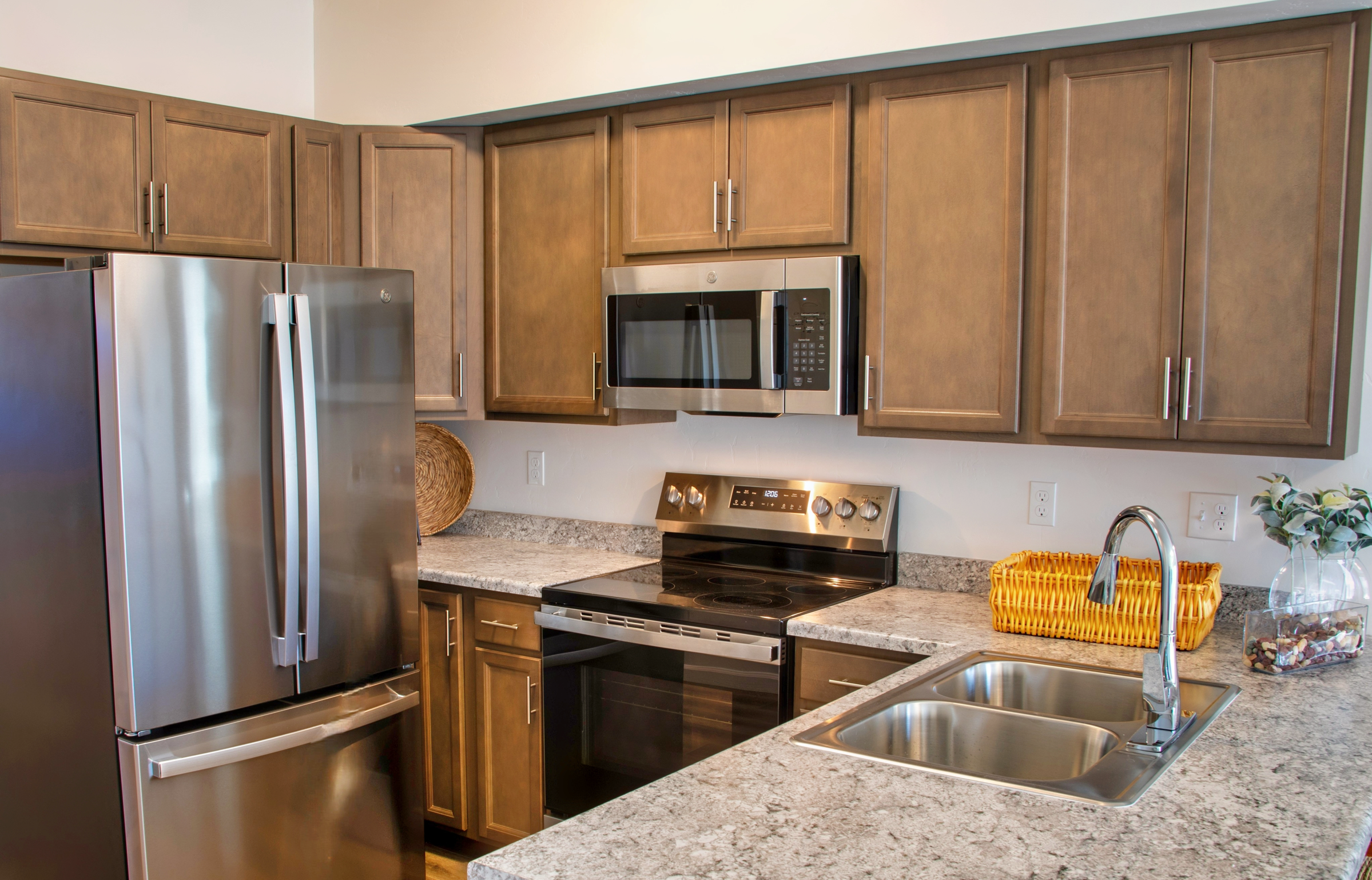 A modern kitchen featuring wooden cabinets, stainless steel appliances including a refrigerator and microwave, granite countertops, and a double sink. A woven basket sits on the counter, adding a touch of warmth to the contemporary design.