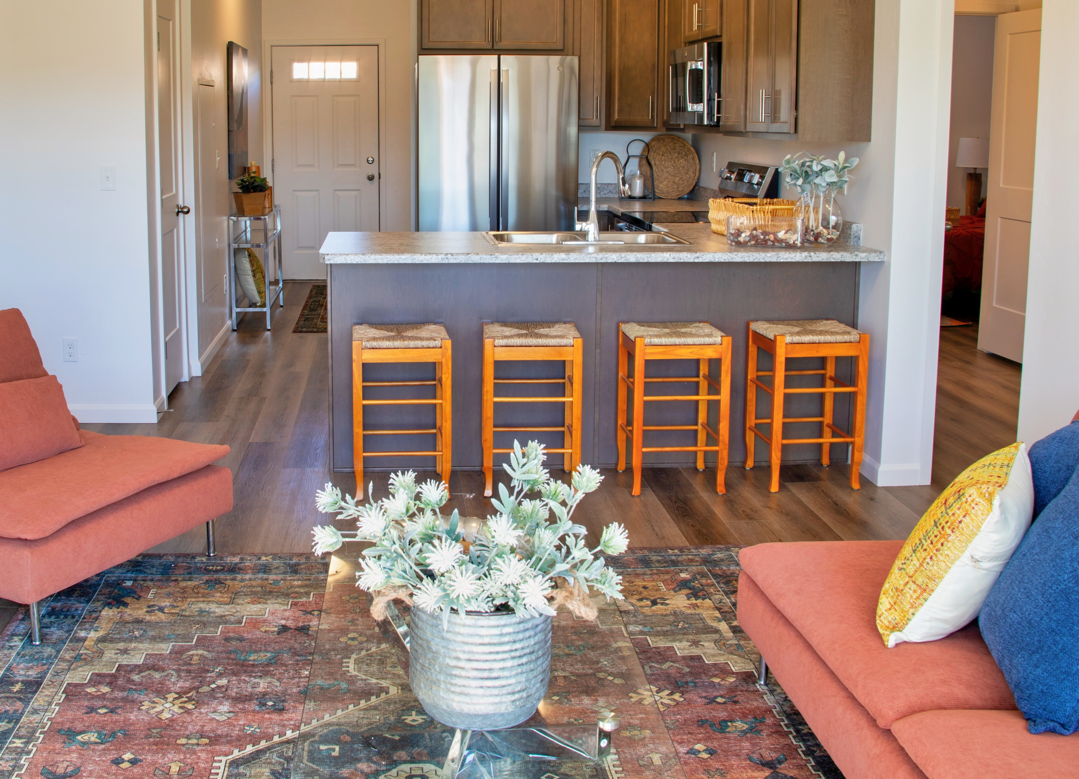 A modern living space featuring a kitchen with stainless steel appliances, a granite countertop, and wooden cabinetry. In the foreground, there are two orange sofas with decorative pillows and a metal planter filled with greenery on a patterned rug. The room has an inviting and stylish atmosphere.
