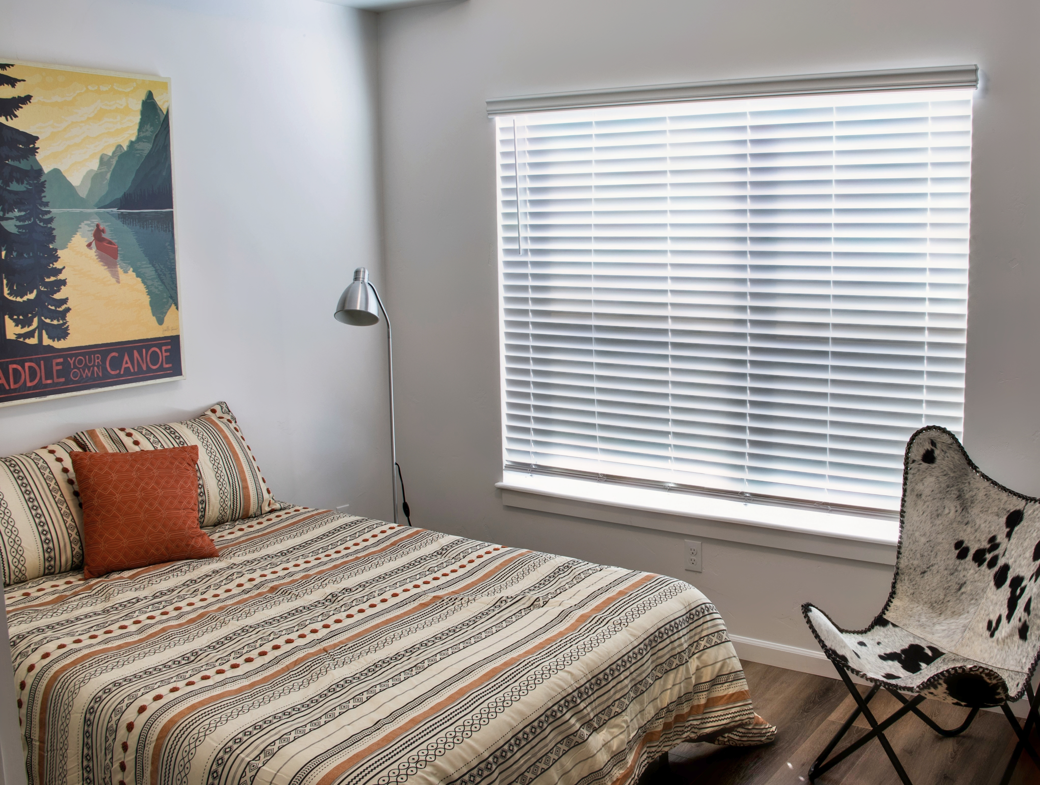 A modern bedroom featuring a striped bedspread with an orange pillow on a neatly made bed. A wooden floor complements the space, which has a large window covered with white blinds. A sleek floor lamp beside the bed provides light, and a chic chair with a cow print design adds a stylish touch. An inspiring nature-themed poster hangs on the wall.