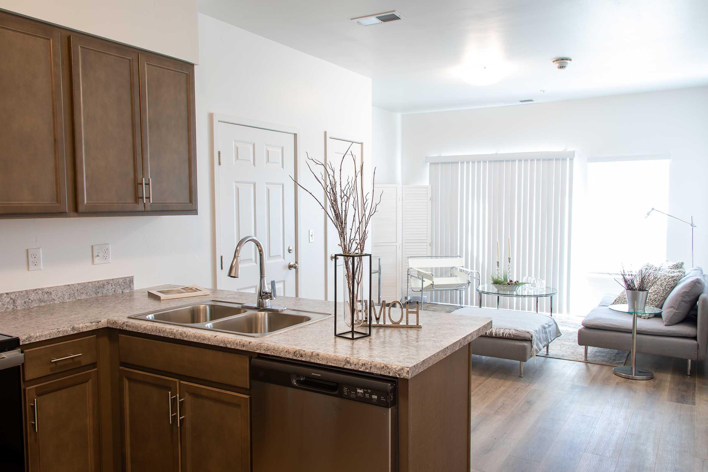 Modern kitchen with dark wood cabinetry, granite countertop, and stainless steel appliances. A large window provides natural light. In the foreground, a decorative vase with branches is placed on the counter. An adjacent living area features a sofa and sliding glass doors, creating a spacious, inviting atmosphere.