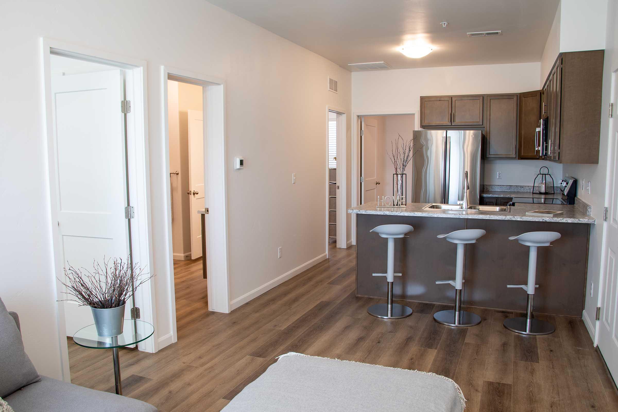 Modern open-concept living space featuring a kitchen with stainless steel appliances and a breakfast bar with three stools. Light wood flooring complements neutral wall colors. A cozy seating area is visible in the foreground, and two doorways lead to additional rooms.