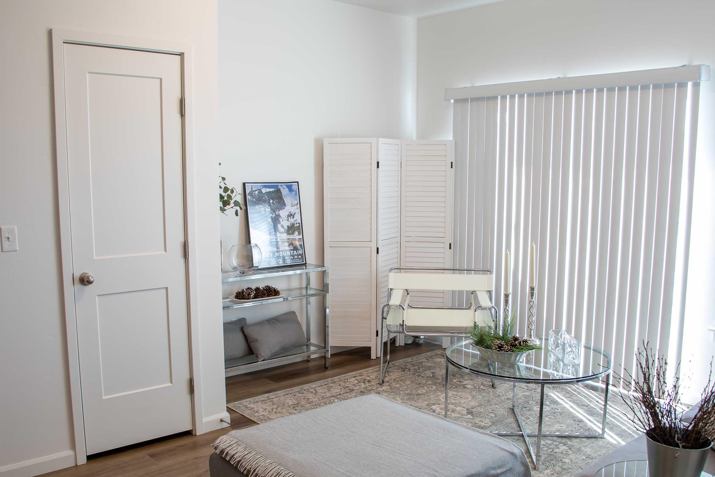 A bright, modern living room with a white door, large windows covered by vertical blinds, and a sleek glass table. A white folding screen is in the corner, alongside a decorative print and a small arrangement of plants. Soft furnishings and a patterned rug add a warm touch to the minimalist decor.