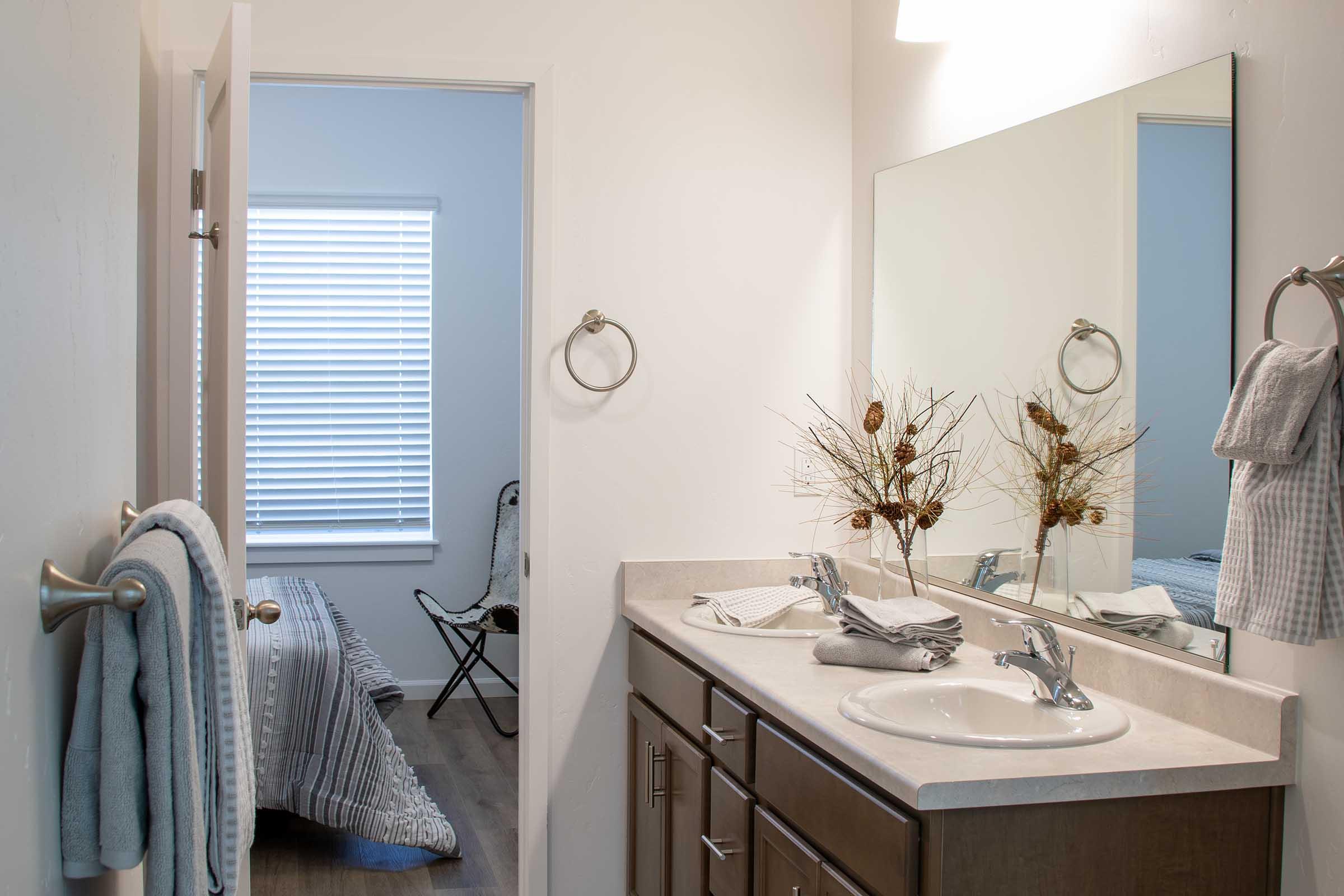 A bright, modern bathroom featuring a white sink with silver fixtures, a large mirror above it, and decorative dried plants. A towel hangs on a ring next to the sink, and another towel is draped over a nearby bar. A doorway leads to a softly lit bedroom with a chair and window covered by blinds.