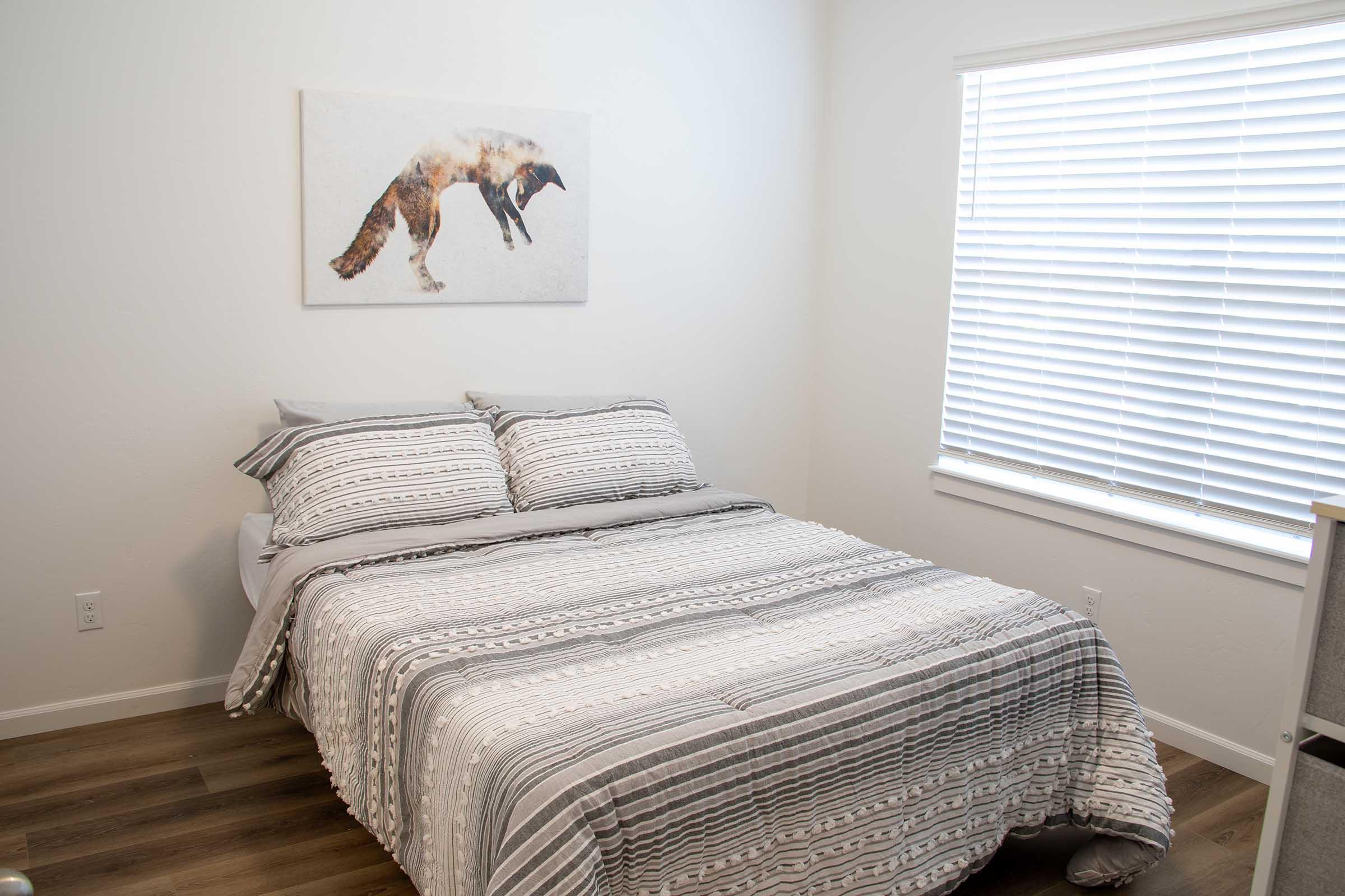 A cozy bedroom featuring a neatly made bed with gray and white striped bedding. A piece of wall art depicting a fox hangs above the bed. The room has white walls, a window with blinds, and light wooden flooring, creating a bright and inviting atmosphere.