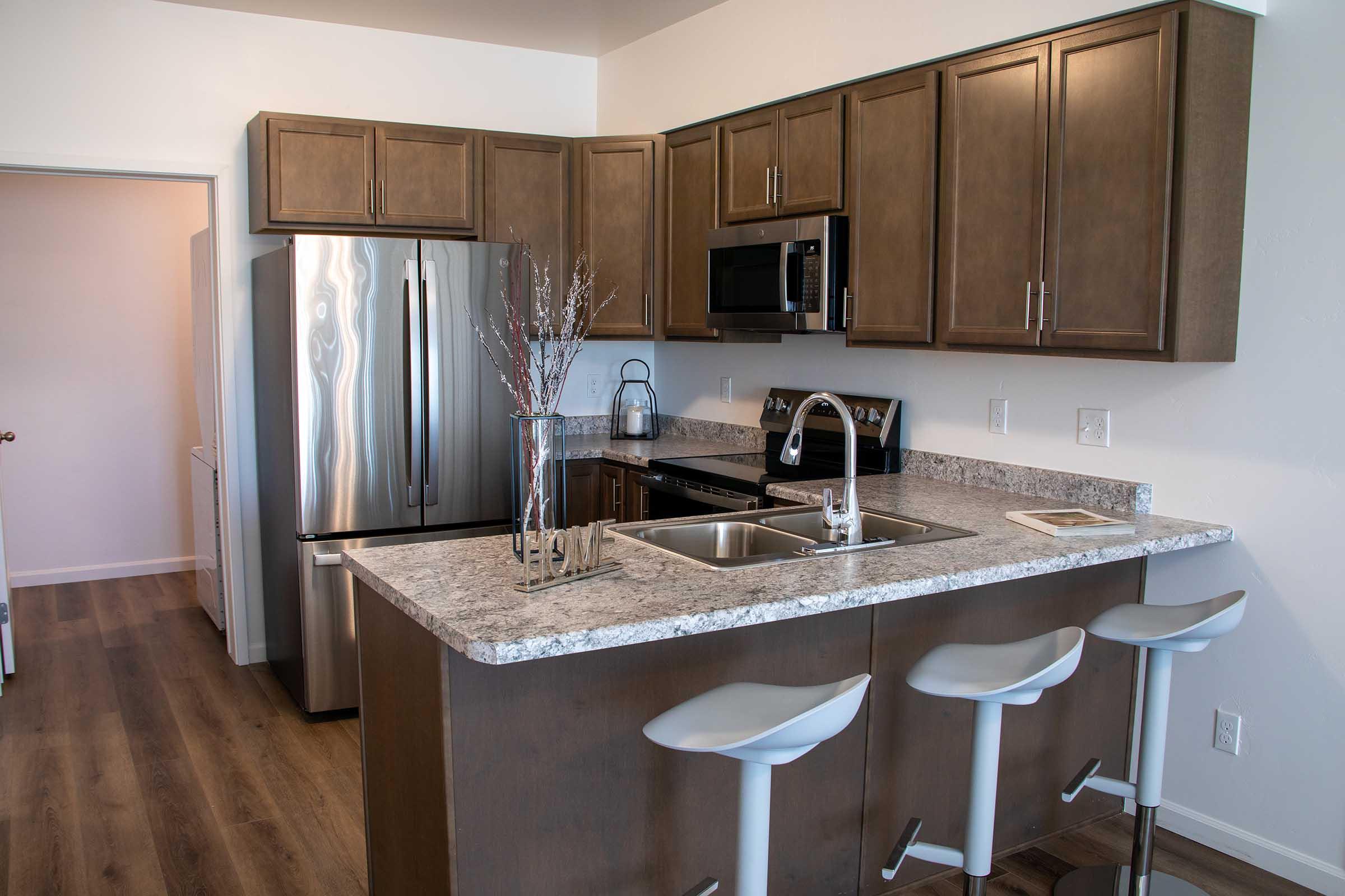 Modern kitchen featuring wooden cabinets, stainless steel appliances, and a granite countertop with three white bar stools. A small decorative arrangement is placed by the sink, and the space has a clean, contemporary aesthetic with warm lighting and stylish finishes.
