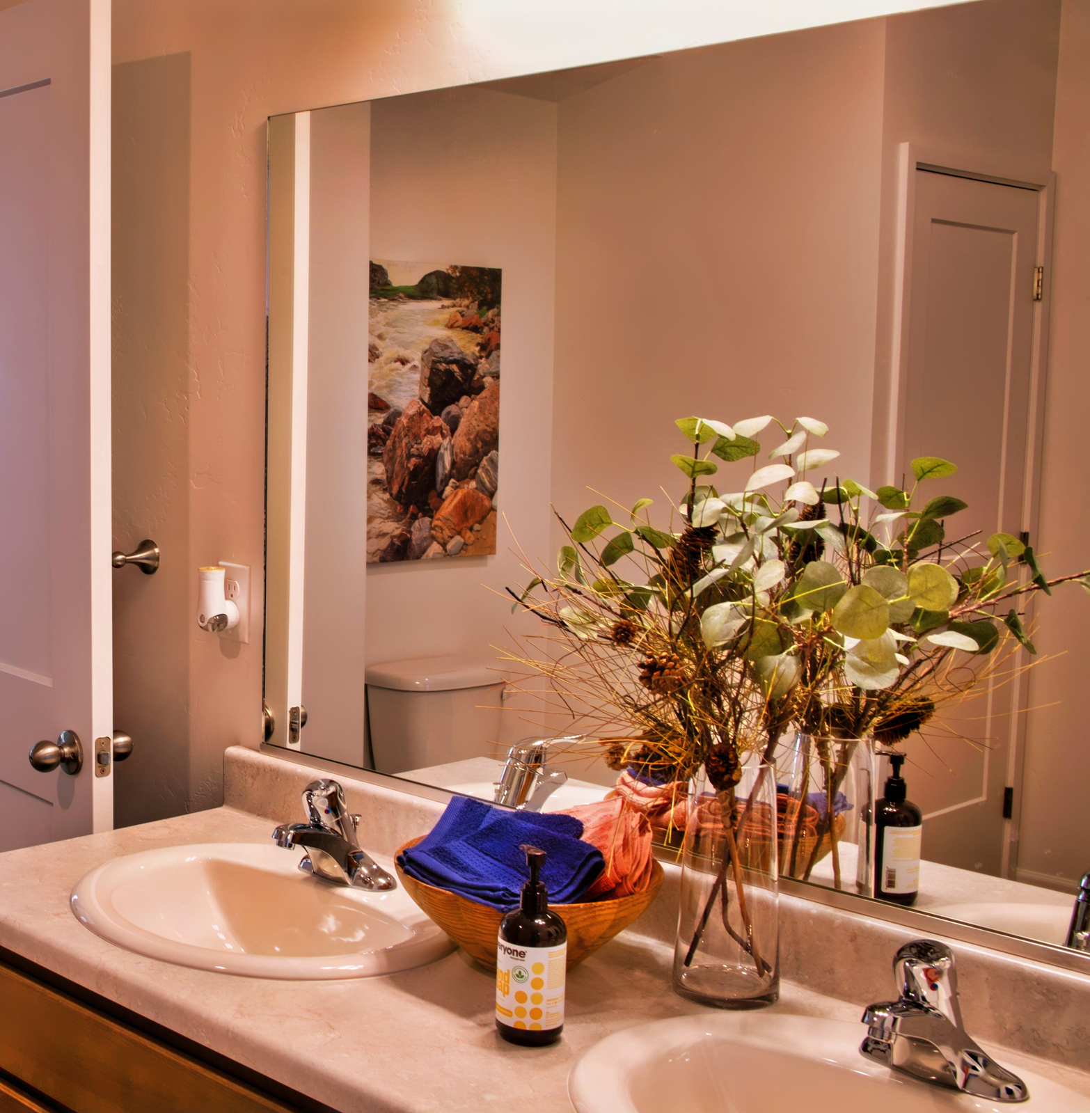 A well-lit bathroom featuring a double sink with a neutral countertop. A decorative vase with eucalyptus sits on the counter alongside a woven basket containing a blue towel. A mirror reflects a colorful nature-themed painting on the wall, and a white door and toilet are visible in the background.