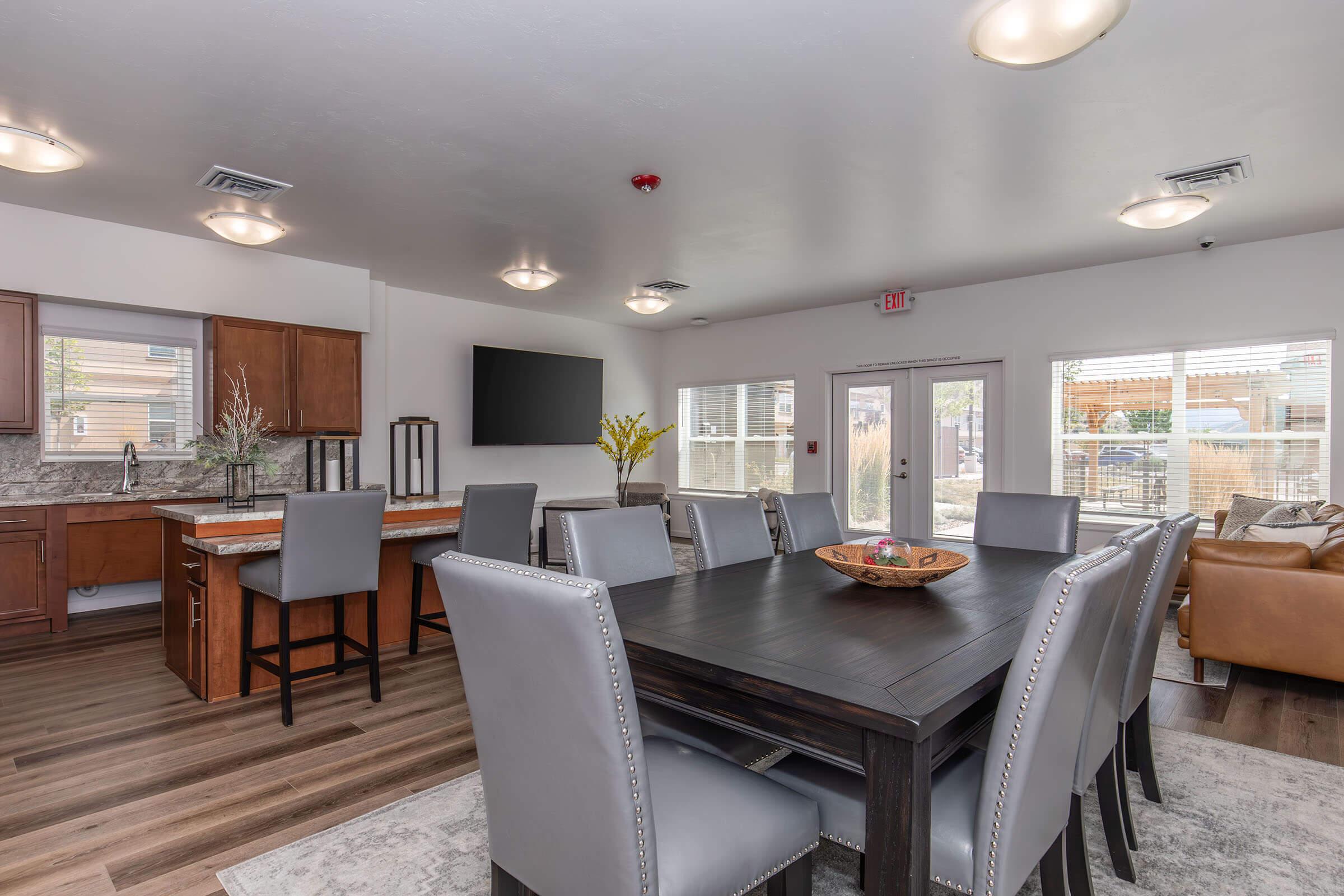 A modern dining area with a large dark wooden table surrounded by gray upholstered chairs. In the background, there is a kitchen with wooden cabinets and a countertop, along with a flat-screen TV mounted on the wall. Large windows allow natural light to fill the space, highlighting the contemporary decor.
