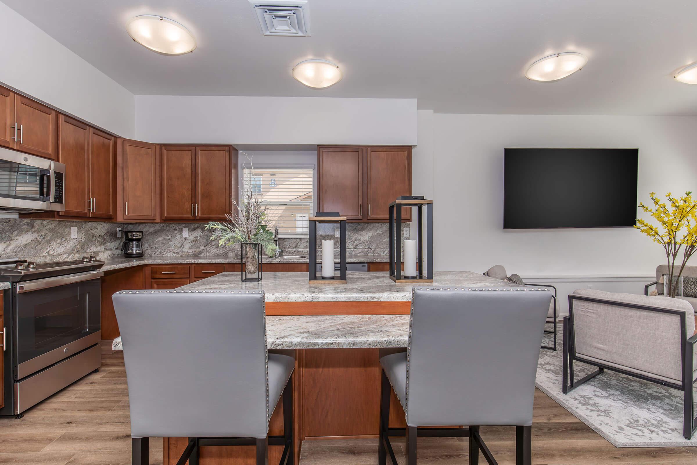 Modern kitchen with wooden cabinets and stainless steel appliances. A breakfast bar with two gray stools is in the foreground. The countertop is made of granite, and there’s a large TV mounted on the wall in the background. Decorative elements include a vase with flowers and a stylish living area adjacent to the kitchen.