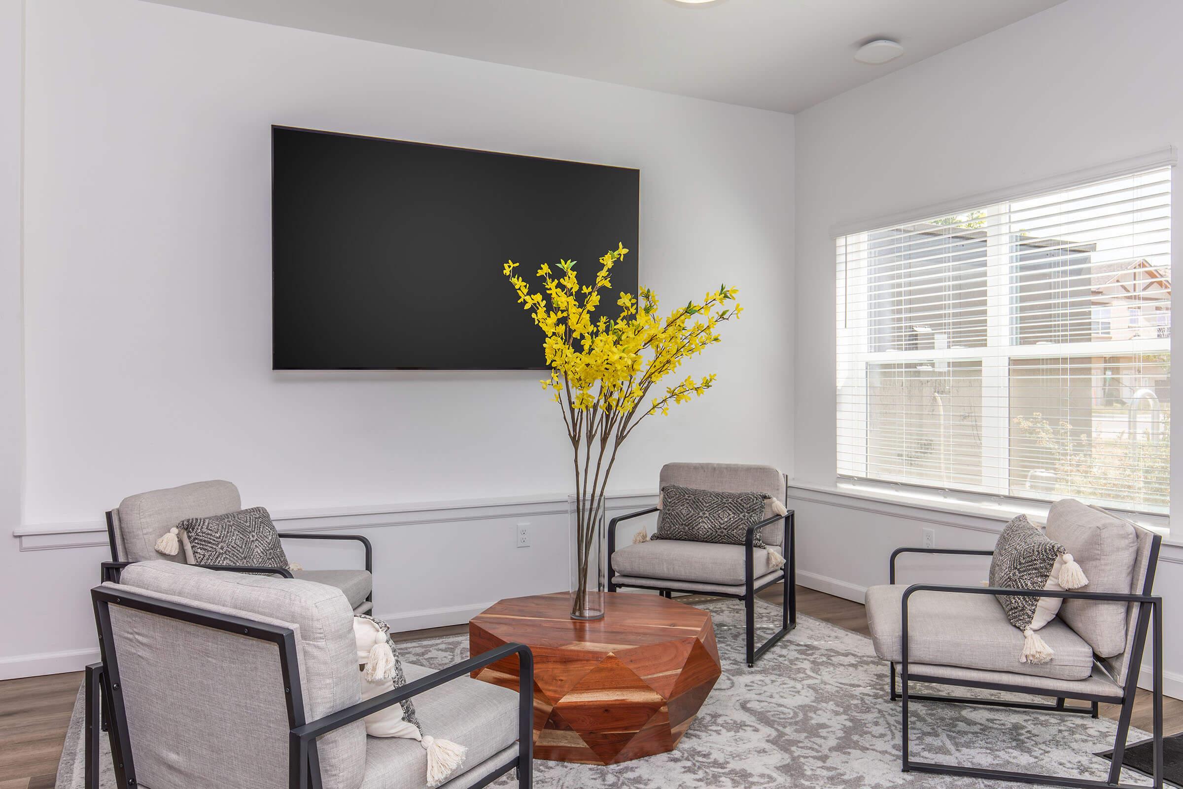 A modern living room featuring a large flat-screen TV mounted on the wall. In the center, a wooden hexagonal coffee table is surrounded by four stylish armchairs. A tall vase with bright yellow flowers adds a pop of color. Natural light floods the space through a nearby window with blinds.