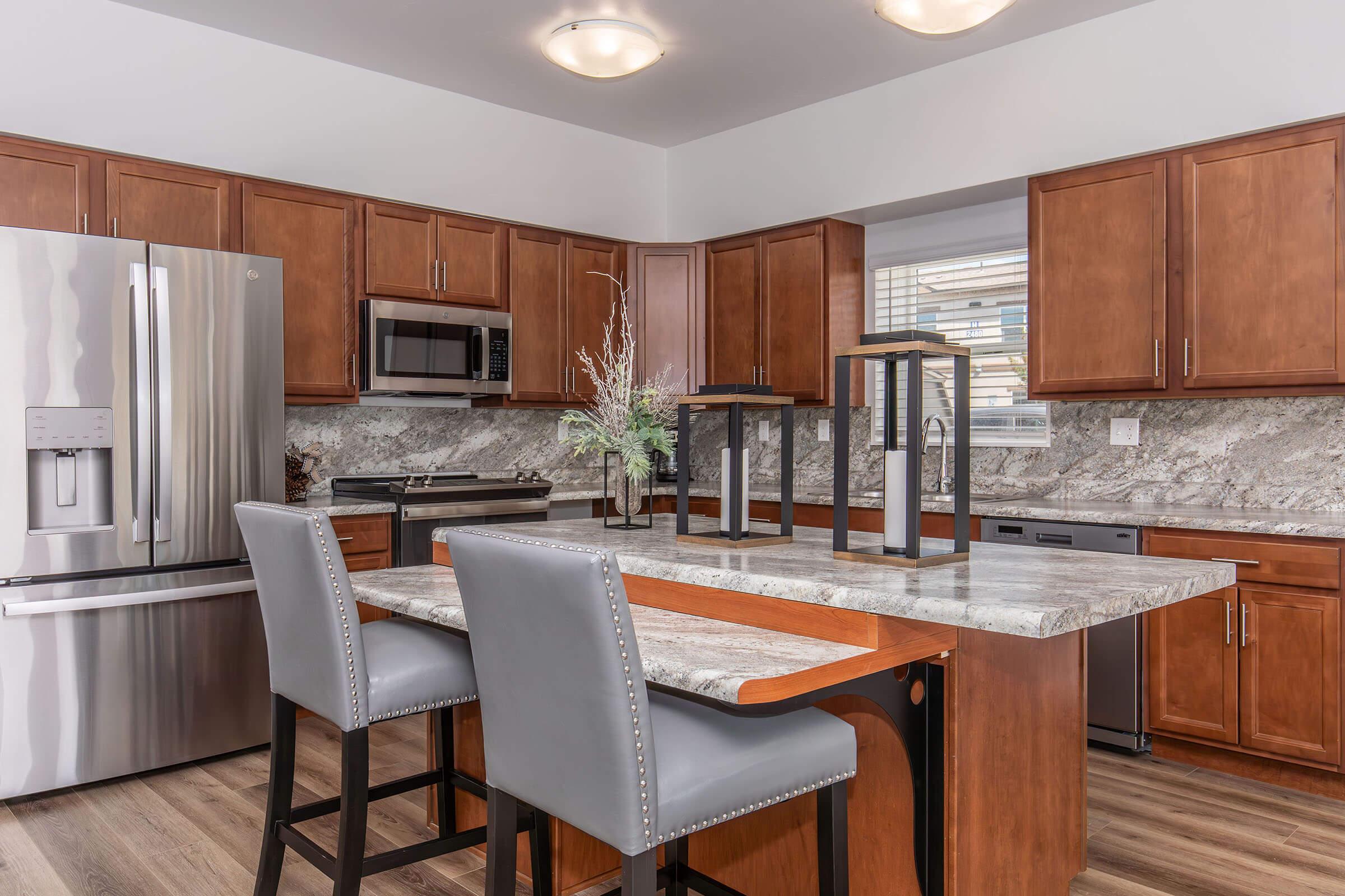 A modern kitchen featuring wooden cabinets, a large island with a marble countertop, and stainless steel appliances, including a refrigerator and microwave. Two elegant gray bar stools are placed at the island, and decorative elements are arranged on the countertop, adding a sophisticated touch to the space.