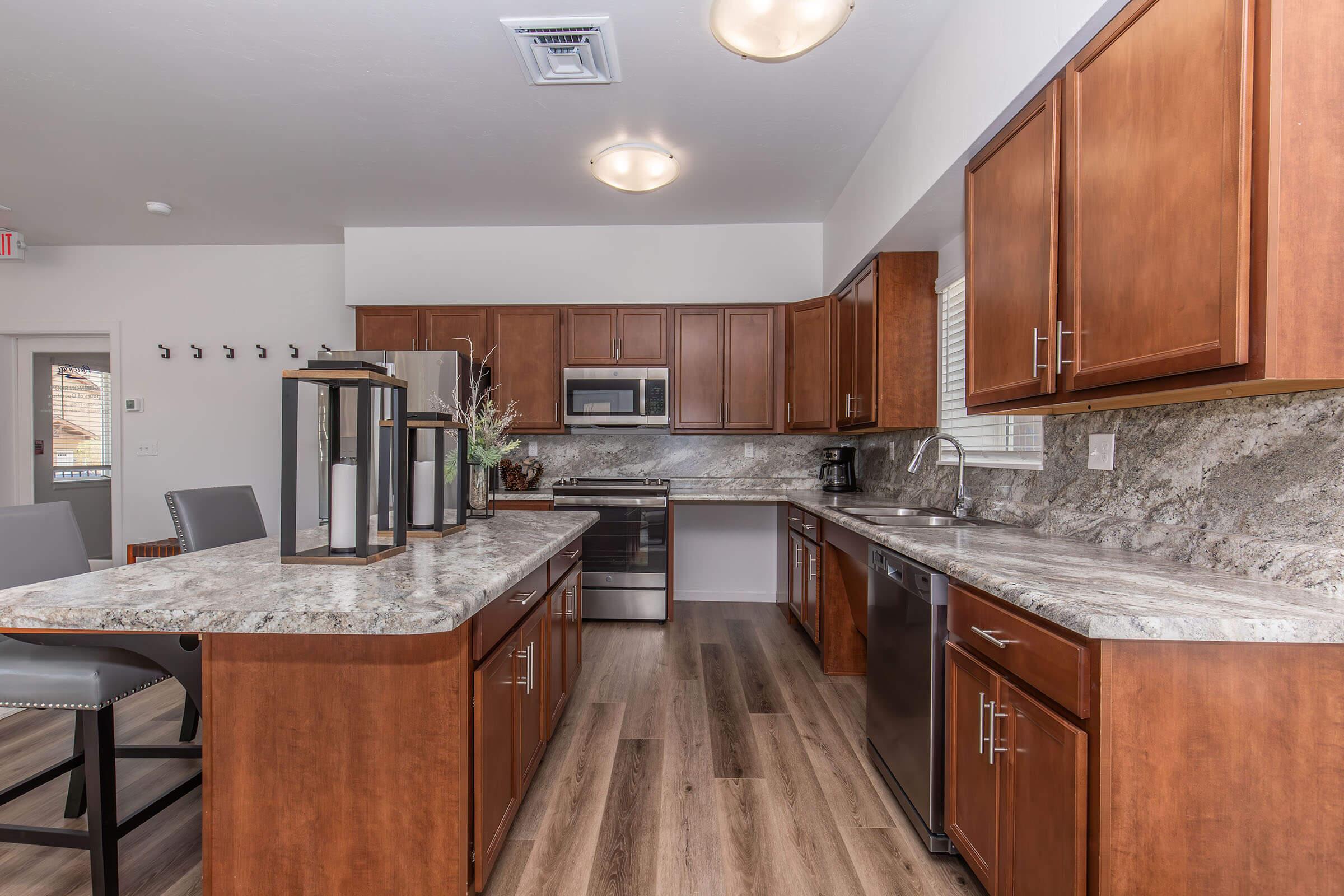 Spacious modern kitchen featuring wooden cabinetry, a large granite countertop with seating, stainless steel appliances including a microwave and dishwasher, and ample natural light through windows. The floor is made of light wood, enhancing the contemporary design of the space.
