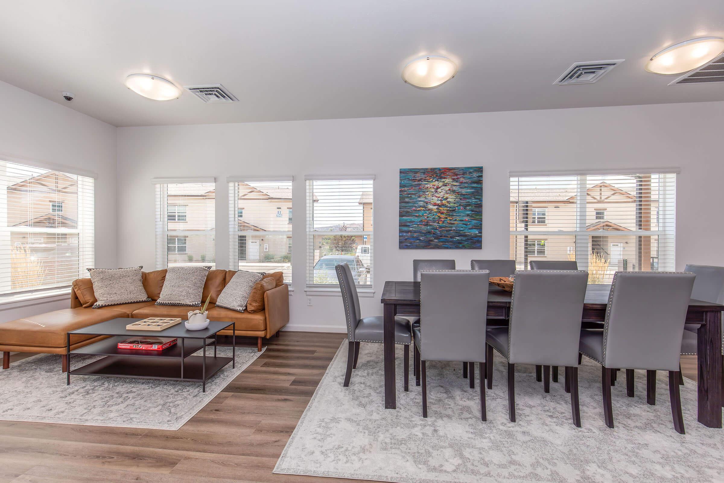 Modern living room and dining area featuring a brown leather sofa, wooden coffee table, and a large dining table with gray chairs. The room has bright natural lighting from large windows overlooking houses outside. A colorful abstract painting adorns the wall, and the flooring is a combination of wood and rug.