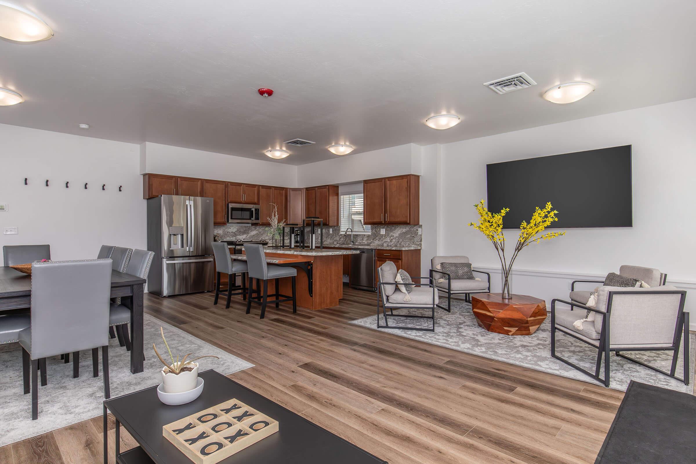 Spacious interior of a modern living area featuring a stylish kitchen with stainless steel appliances, a dining table with chairs, and comfortable seating. The decor includes a coffee table with decorative items, a large wall-mounted TV, and a potted plant, creating a welcoming atmosphere.