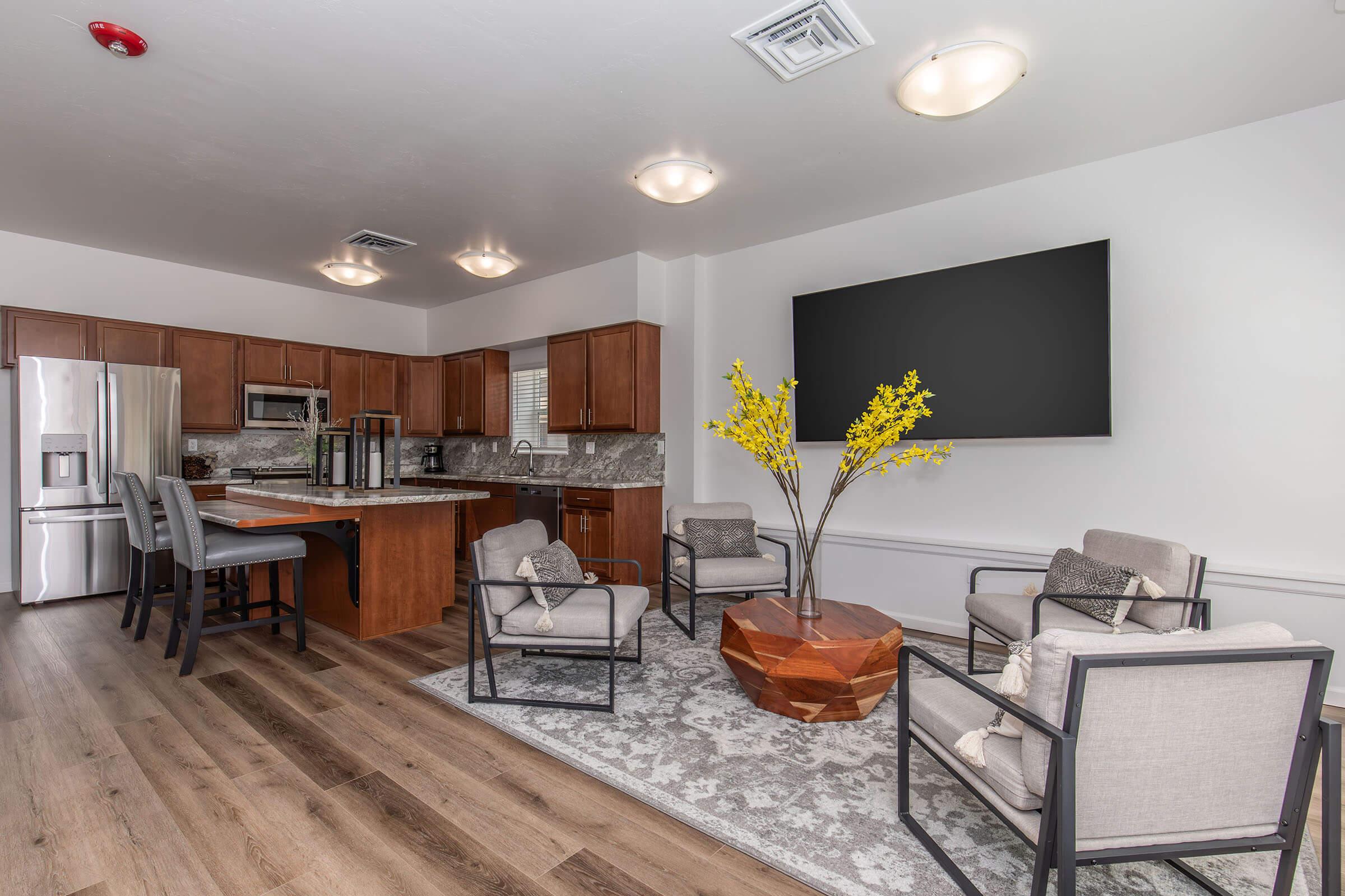 A modern living space featuring a kitchen area with wooden cabinets, stainless steel appliances, and a large flat-screen TV on the wall. Stylish seating is arranged around a geometric coffee table, complemented by decorative yellow flowers in a vase. The flooring is hardwood, and a light rug adds comfort to the space.