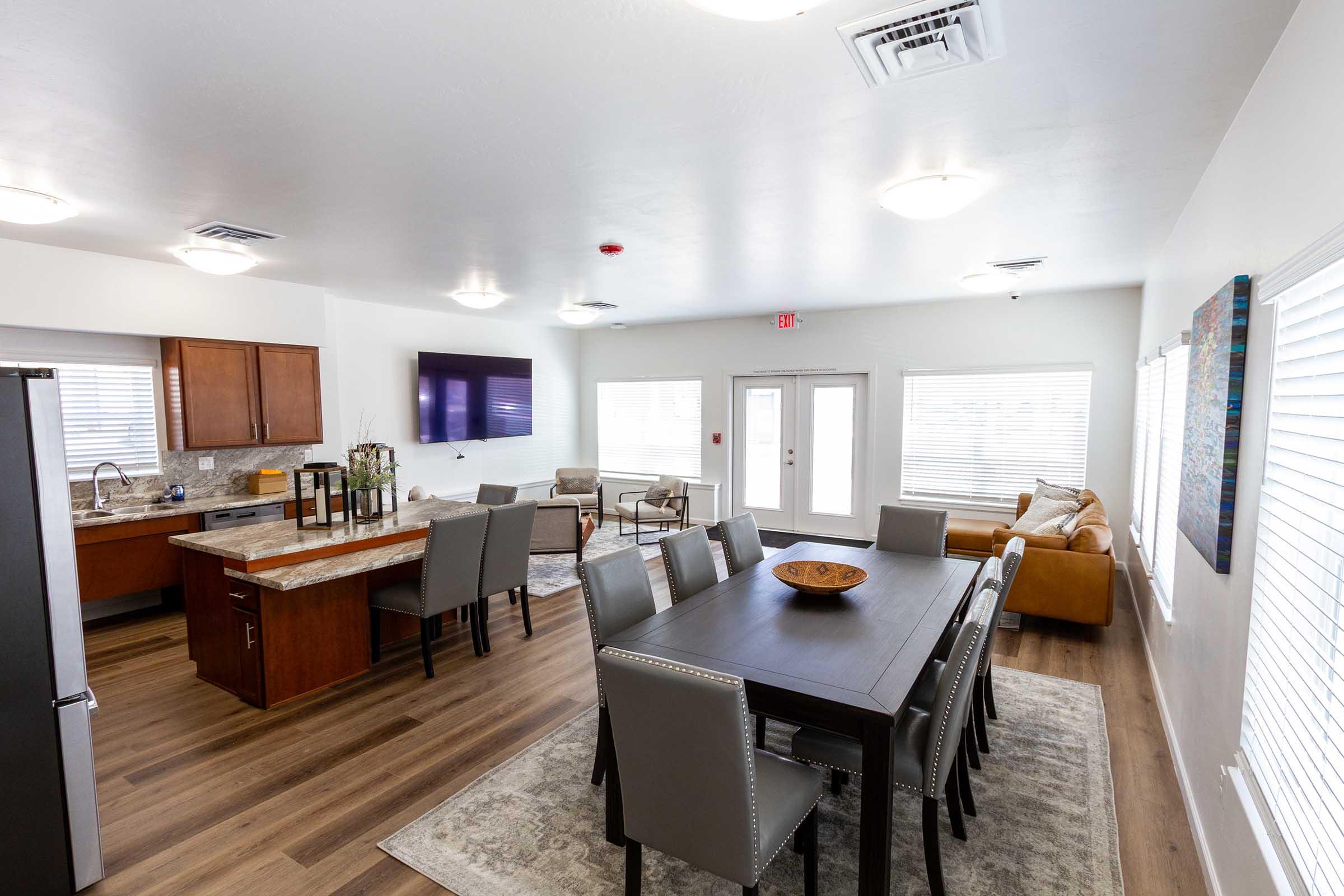 A modern open-concept dining and living area featuring a dining table with grey chairs, a cozy sofa, and a kitchen with wood cabinets and stainless steel appliances. Large windows allow natural light, and there's a flat-screen TV mounted on the wall. Soft, neutral-toned decor enhances the spacious feel of the room.