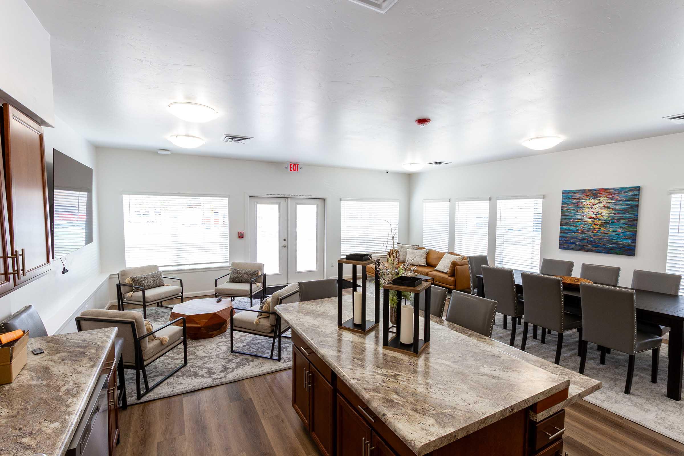A spacious, modern common area featuring a kitchen island with a stone countertop, seating arrangements including couches and dining chairs, large windows providing natural light, and decorative artwork on the wall. The overall atmosphere is welcoming and well-lit.