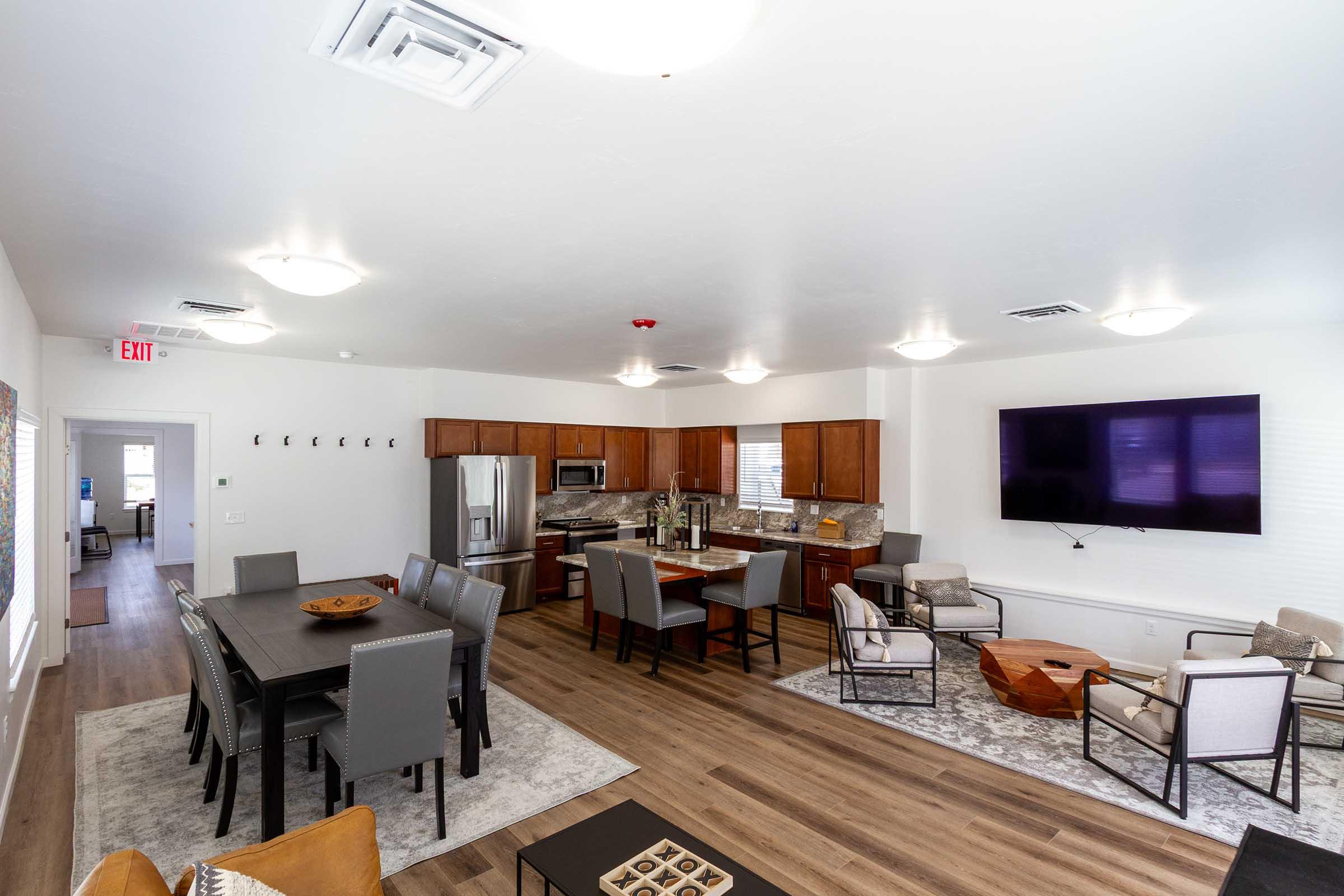 A spacious modern living area featuring a dining table with gray chairs, a cozy seating arrangement with beige armchairs, and a large flat-screen TV mounted on the wall. The kitchen area is equipped with stainless steel appliances and wooden cabinets, all illuminated by overhead lighting in a bright, open layout.