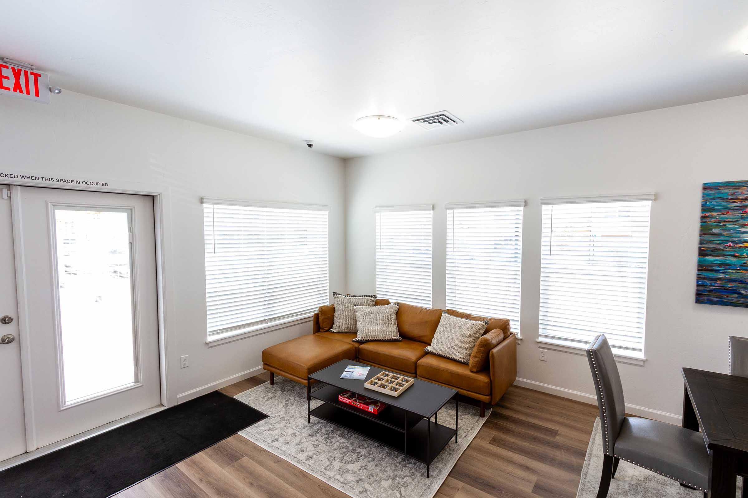 A cozy living room with a brown leather sofa, large windows with white blinds, and a small coffee table. The space features light-colored walls and a dark rug, along with a dining area that includes a gray chair and a wall-mounted artwork. A clear exit sign is visible on the wall.
