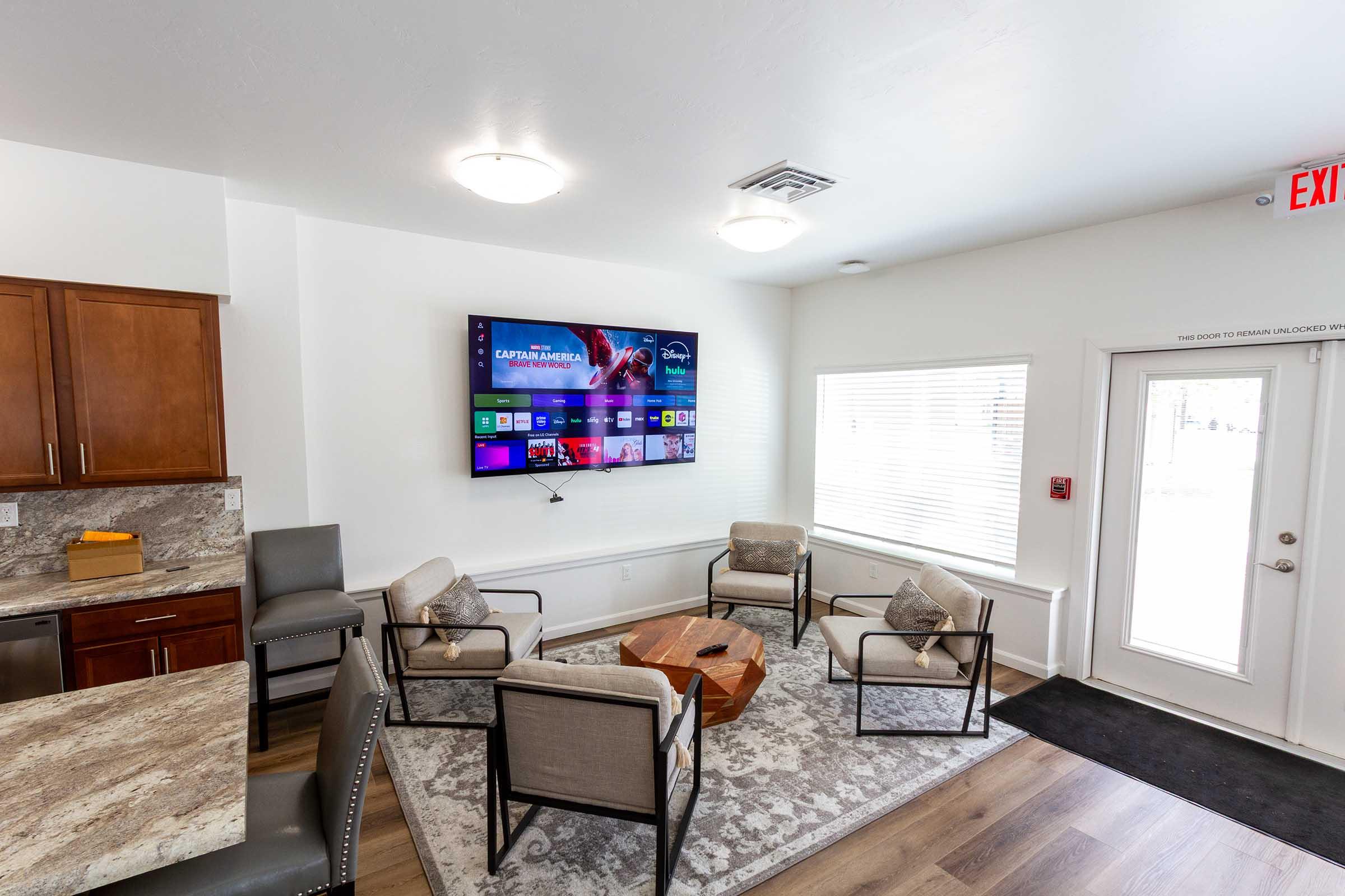 A modern lounge area featuring a large flat-screen TV mounted on a white wall, surrounded by four stylish armchairs arranged around a wooden coffee table. A small kitchenette with dark cabinets is visible in the background, and a door provides access to the outside. Natural light fills the space through a window.