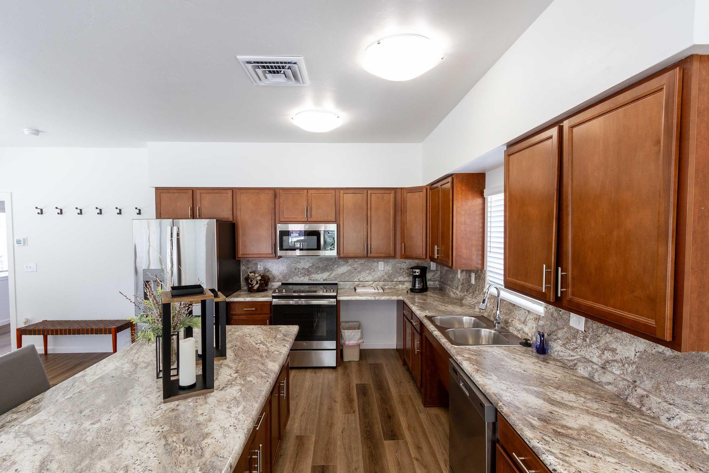 A modern kitchen featuring wooden cabinets, a granite countertop, stainless steel appliances including a refrigerator and microwave, and a double sink. There is a dining table in the foreground and a window with natural light illuminating the space. The floor is finished with wood-like laminate.