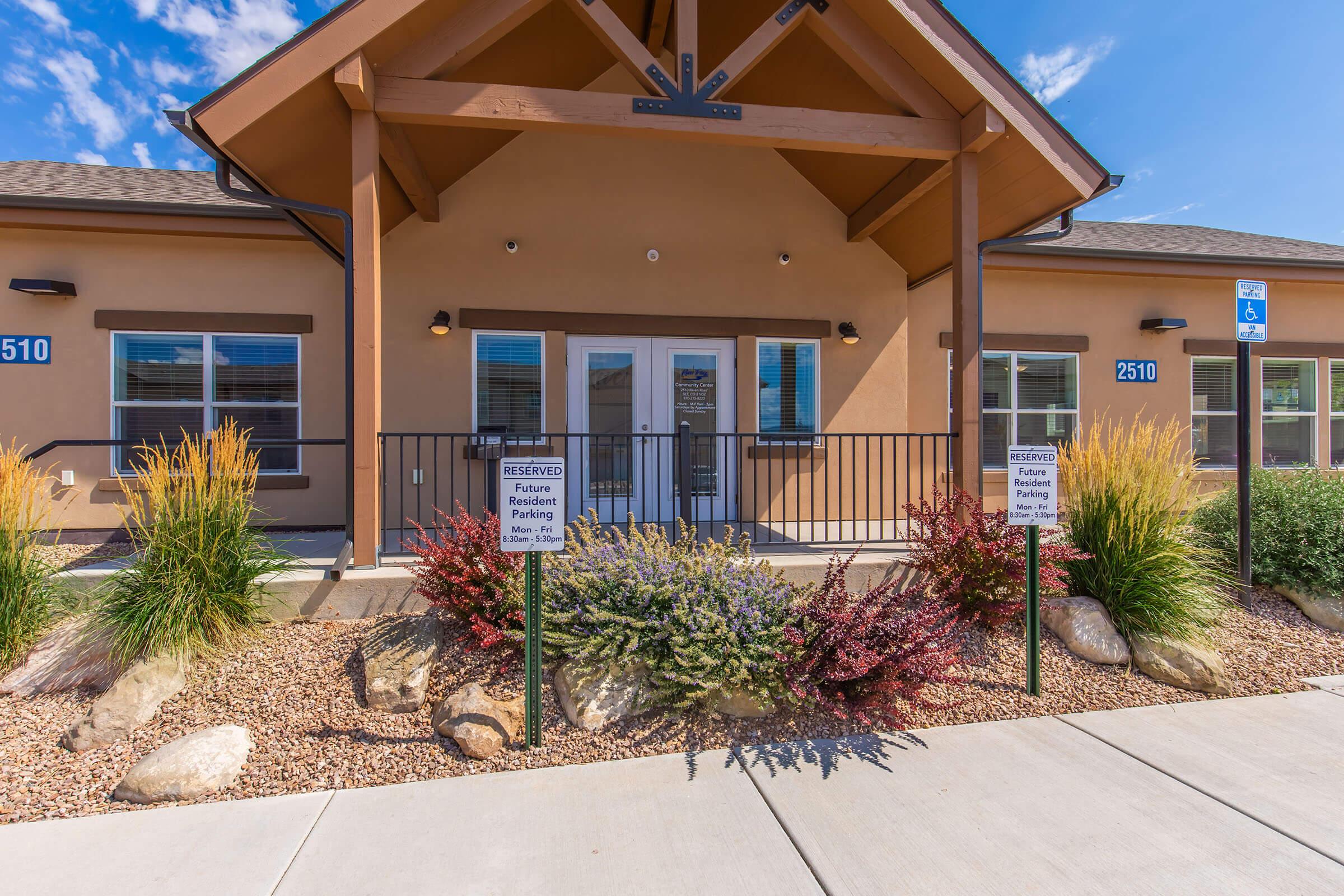 A well-maintained commercial building with a welcoming facade, featuring a covered porch, two front doors, and large windows. The surrounding landscaping includes colorful shrubs and ornamental grasses, with reserved parking signs visible. Clear blue skies and a well-paved walkway enhance the inviting atmosphere.