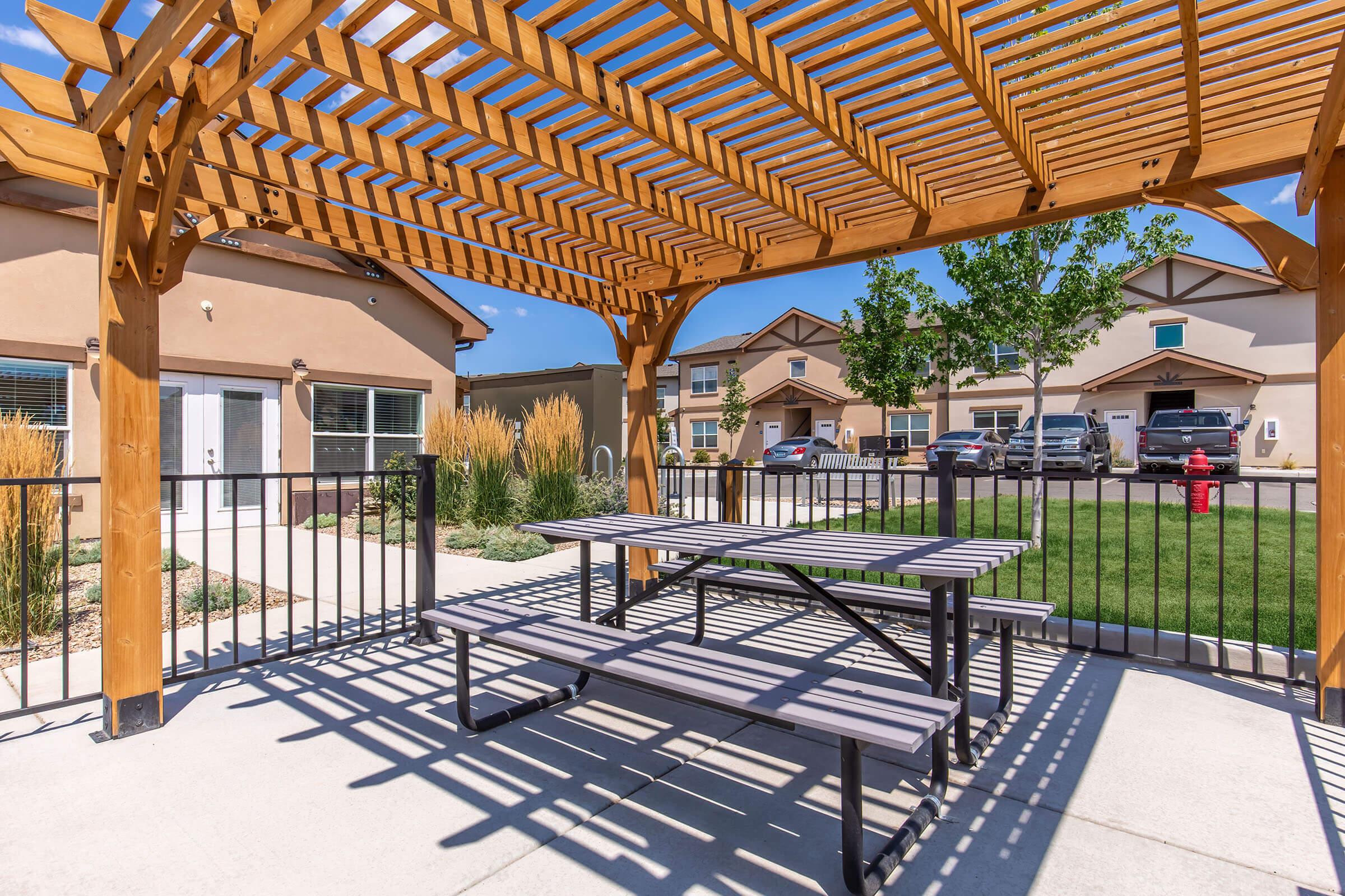 A wooden pergola covers a picnic area with a table and benches, surrounded by a landscaped yard featuring ornamental grasses. Nearby, residential buildings are visible in the background, along with parked cars and a fire hydrant. The scene is bathed in bright sunlight under a clear blue sky.