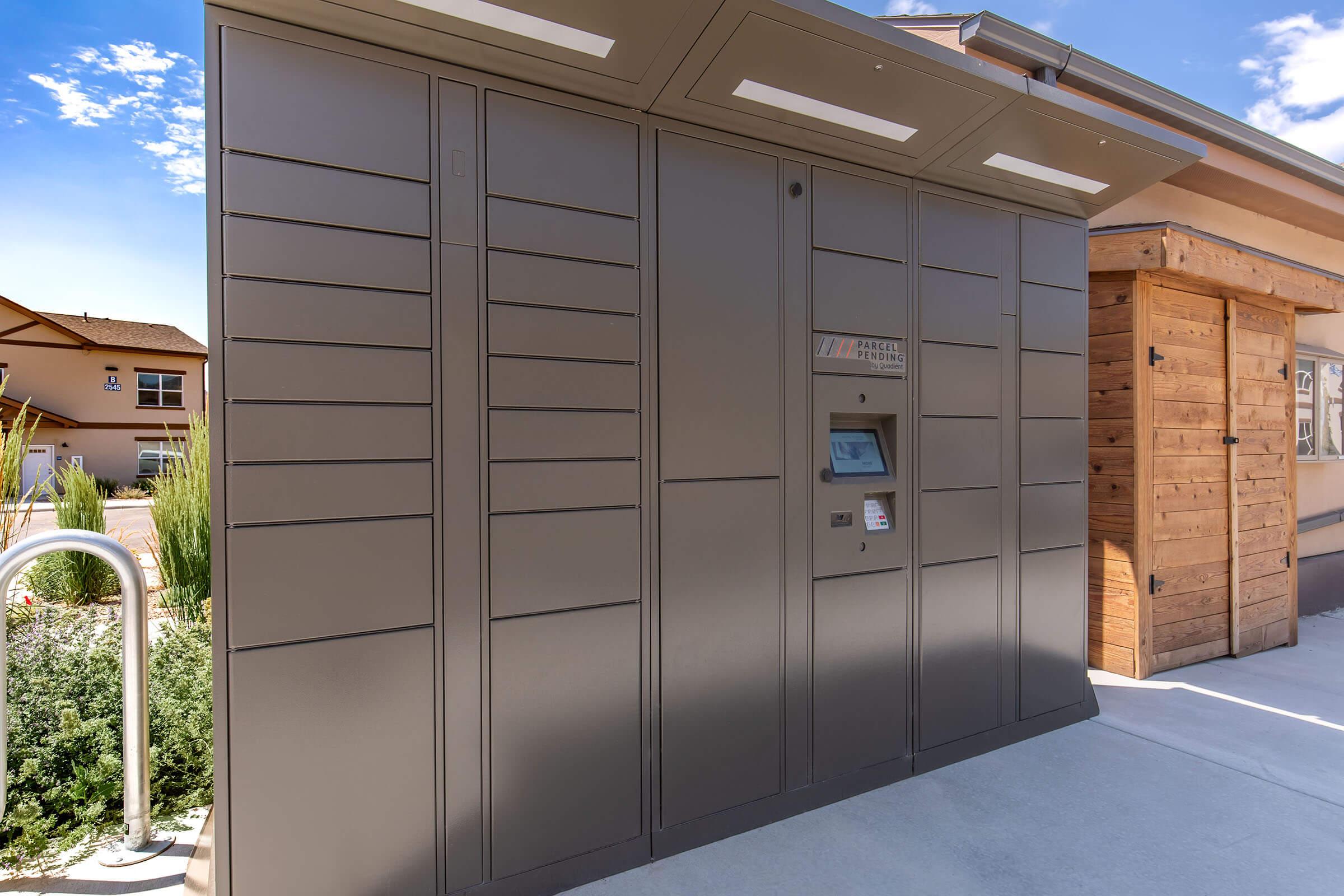 A modern package delivery locker system mounted against a building exterior. The unit features several compartments, a digital touchscreen interface for access, and is situated in a well-maintained outdoor area with greenery and another wooden structure nearby.