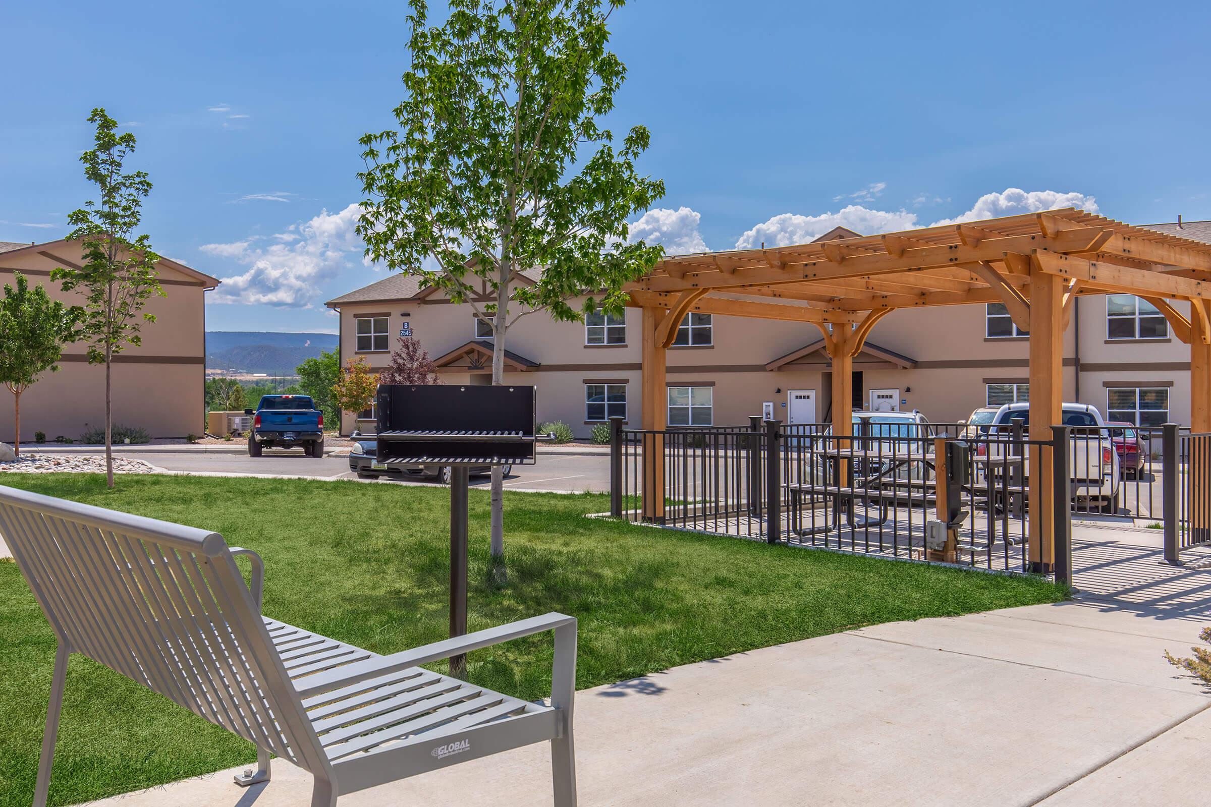 A spacious outdoor area featuring a green lawn, a bench, a barbecue grill, and a pergola. In the background, two buildings can be seen, and there's an area for parking cars. The scene is bright and sunny with a clear blue sky and some clouds.